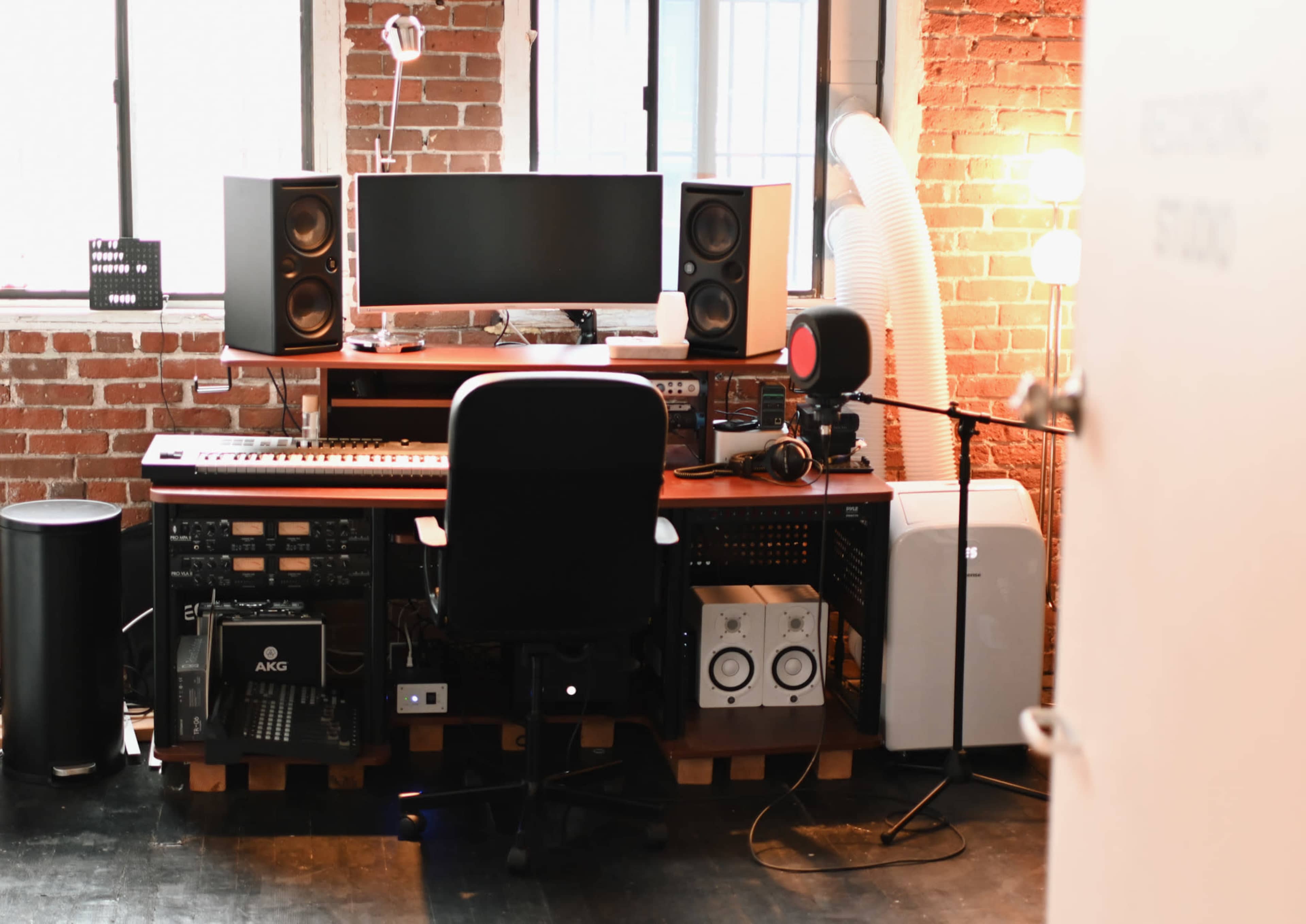 A music production studio setup featuring a desk with a computer monitor, speakers, a keyboard, and various audio equipment against a brick wall.