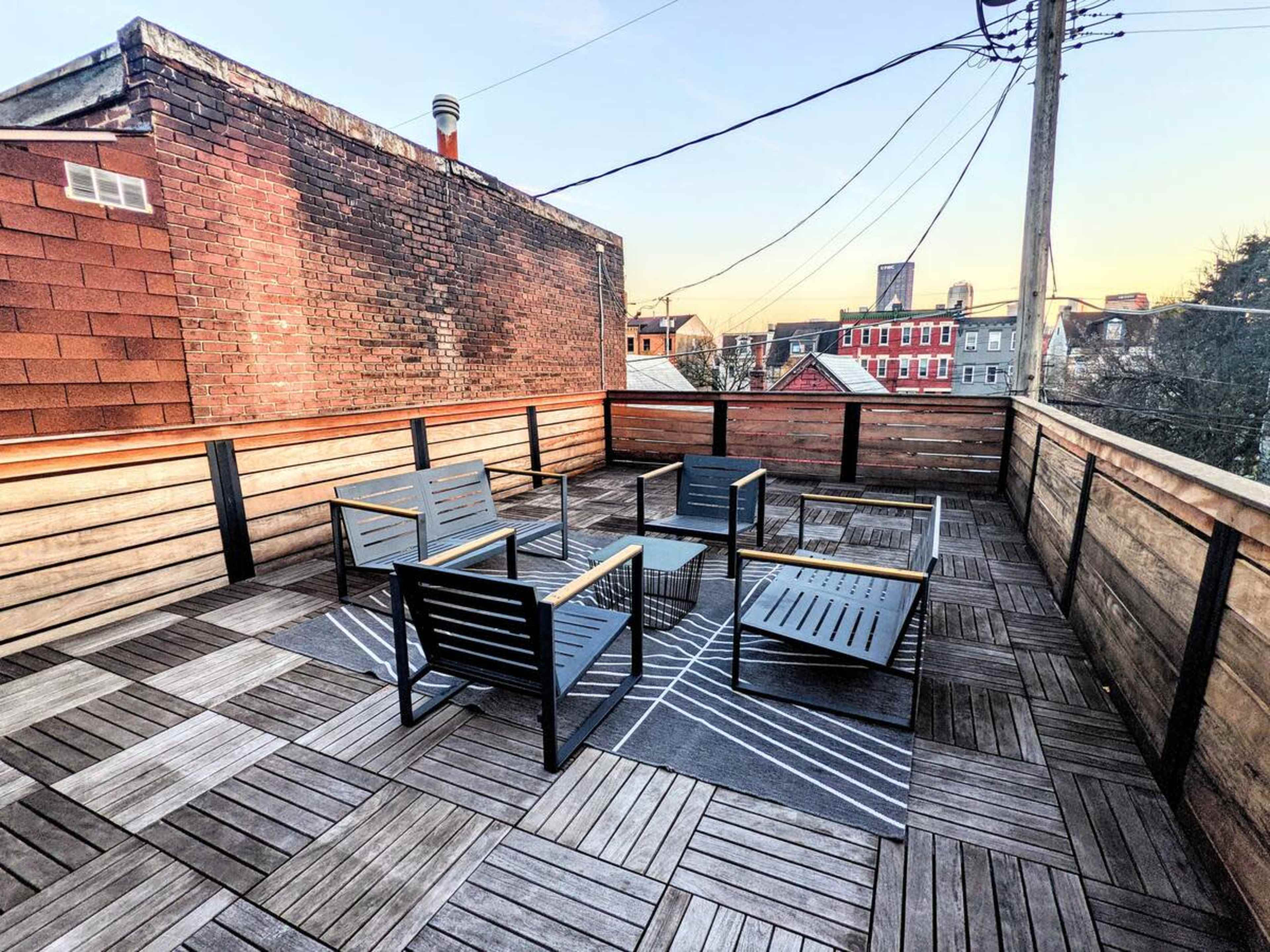 A rooftop terrace with wooden decking and metal-framed seating arranged around a central table, set against a backdrop of urban buildings and power lines.