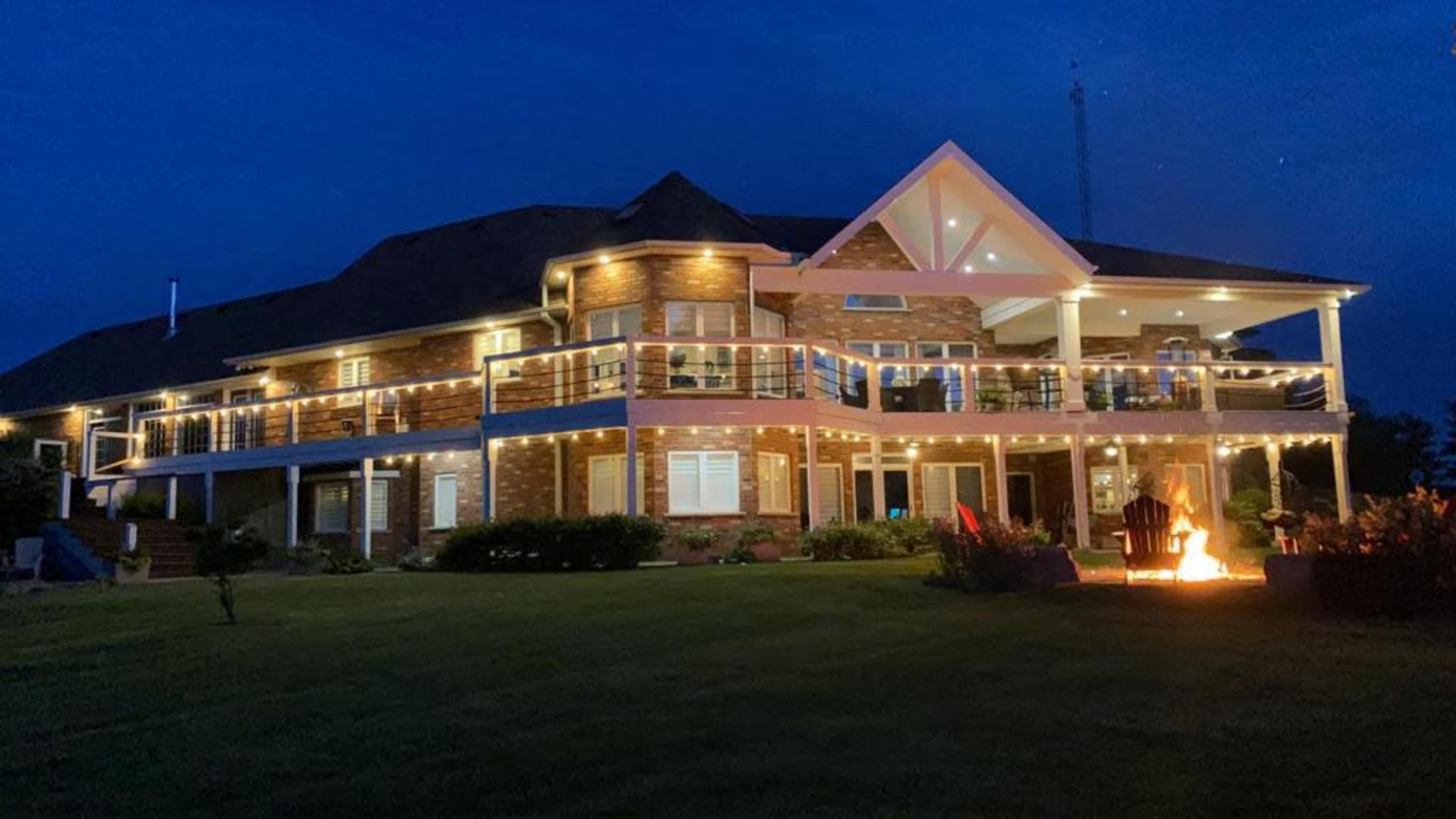A large house with multiple outdoor balconies is illuminated by string lights at night, with a fire pit in the foreground.