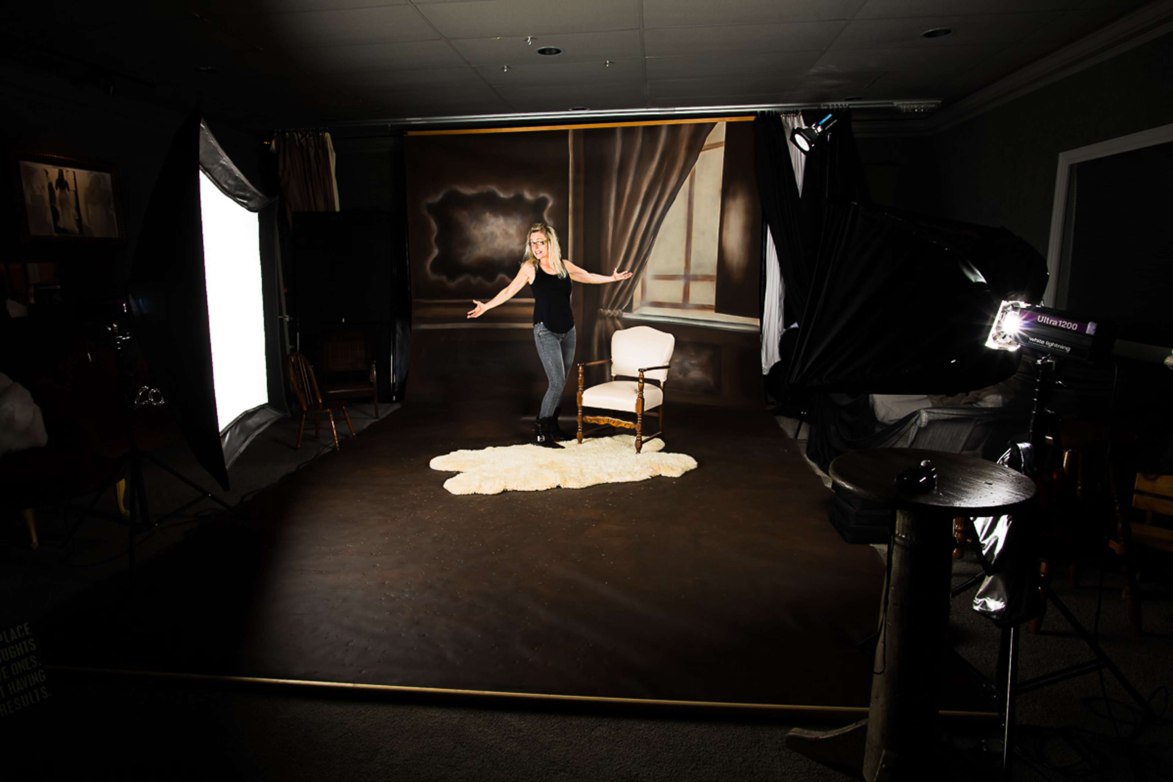 A woman stands beside a white chair on a staged photography set with studio lights and a backdrop depicting a room interior.