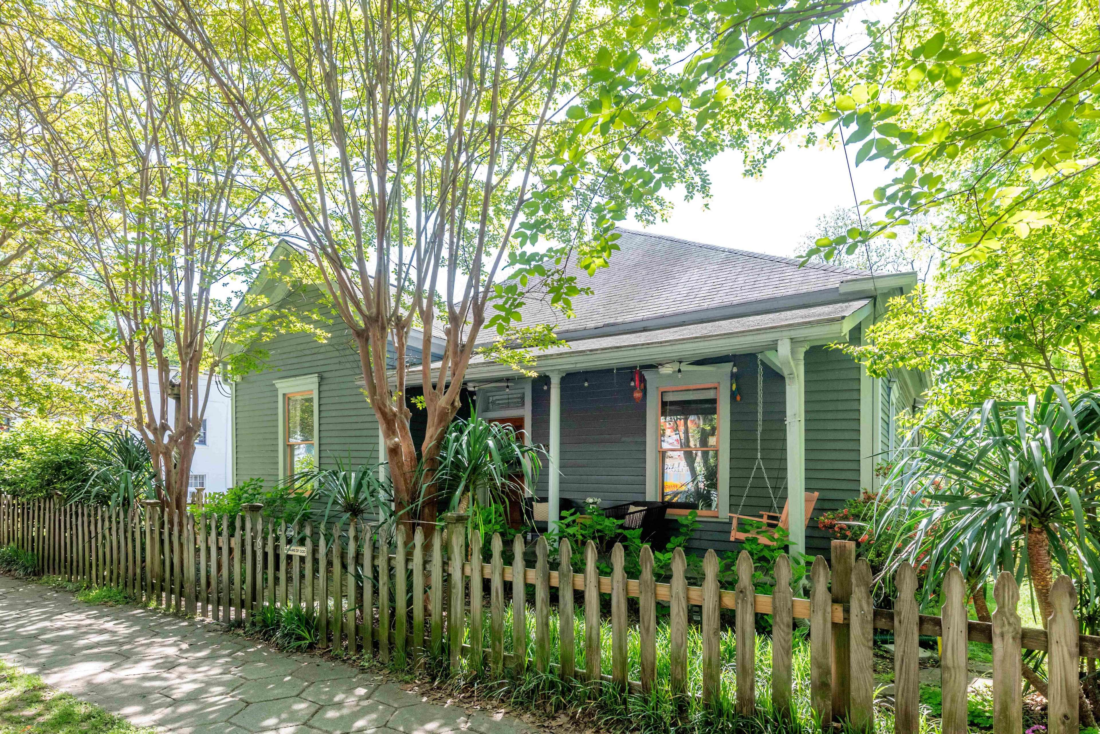 A small, green house with a front porch is surrounded by trees and a wooden fence.