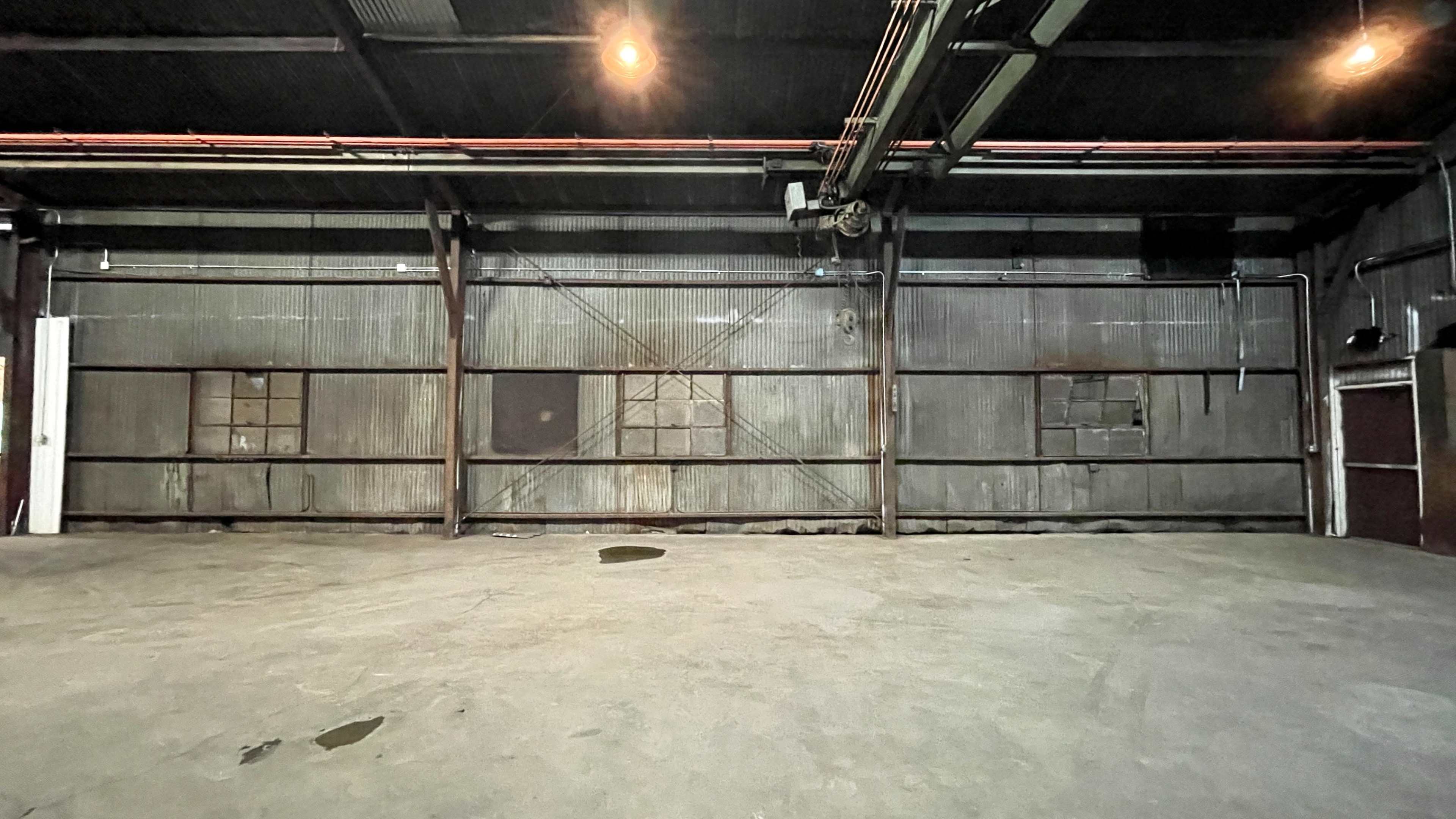 A large, empty industrial space with a concrete floor and metal walls, featuring several windows and overhead lights.