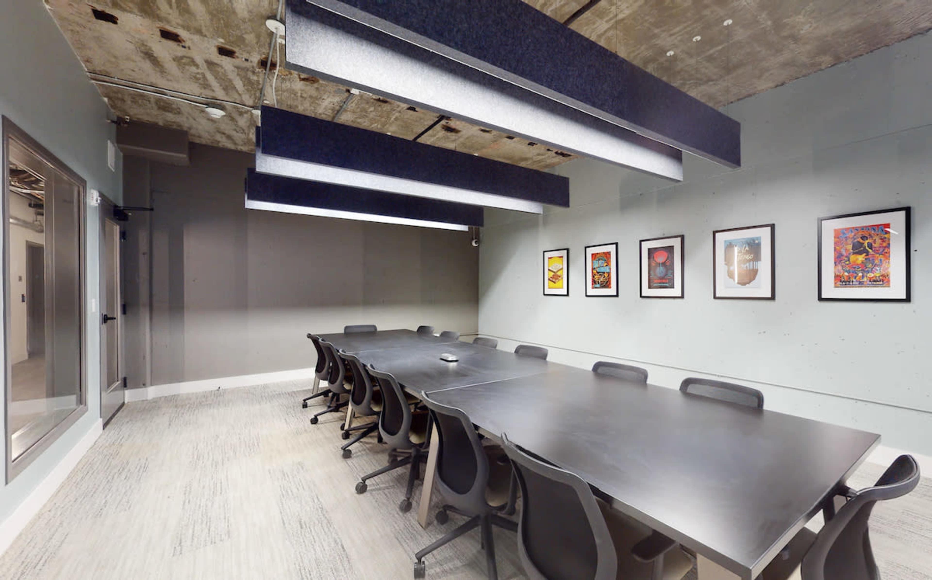 A conference room features a long table surrounded by black chairs, with framed artwork displayed on the walls and modern ceiling lighting.