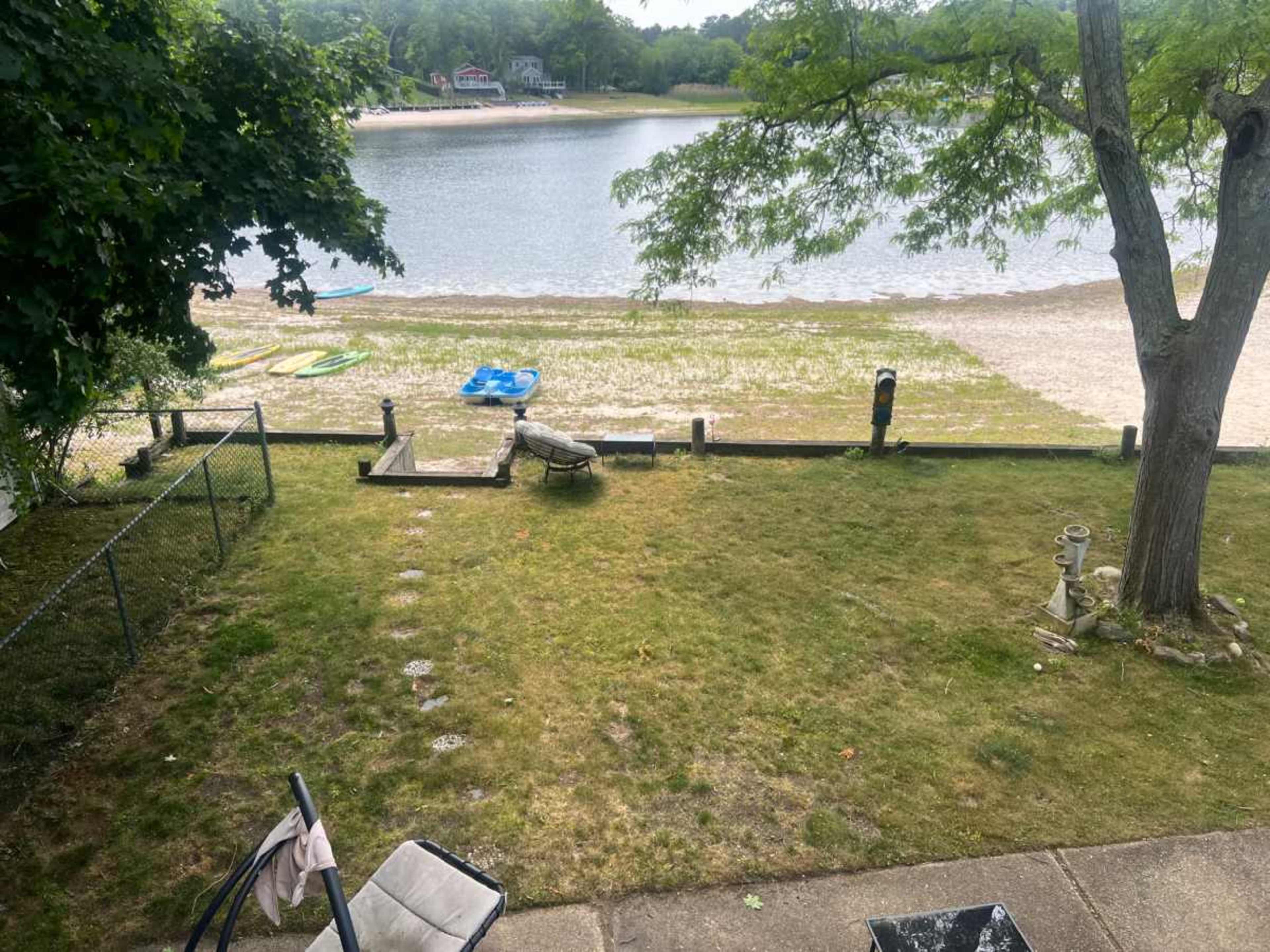 The image shows a grassy area leading to a sandy beach next to a calm body of water, with kayaks and a small boat visible nearby.