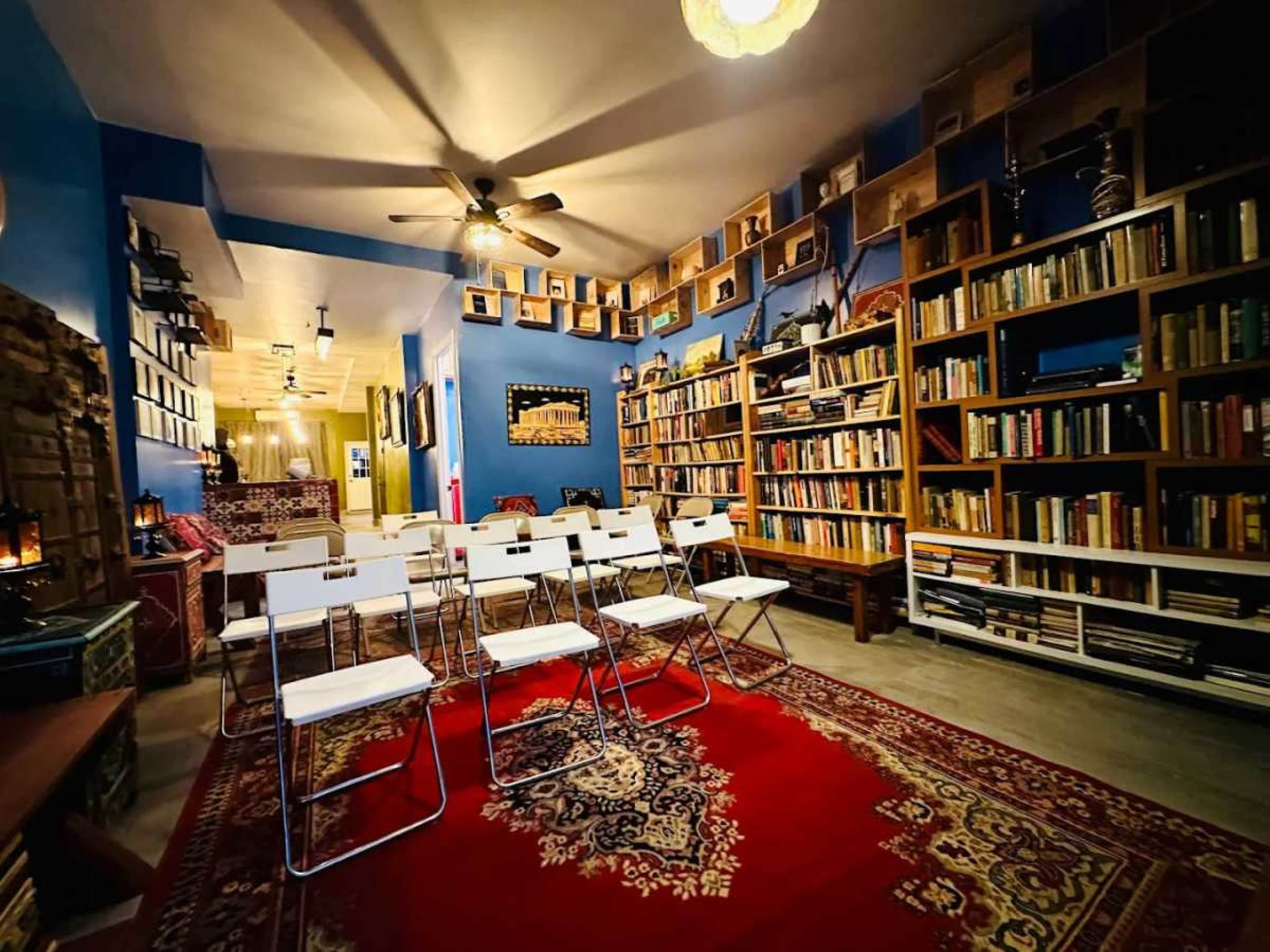 The image depicts a cozy room with bookshelves lining the walls, a patterned rug on the floor, and several white folding chairs arranged in a semi-circle.