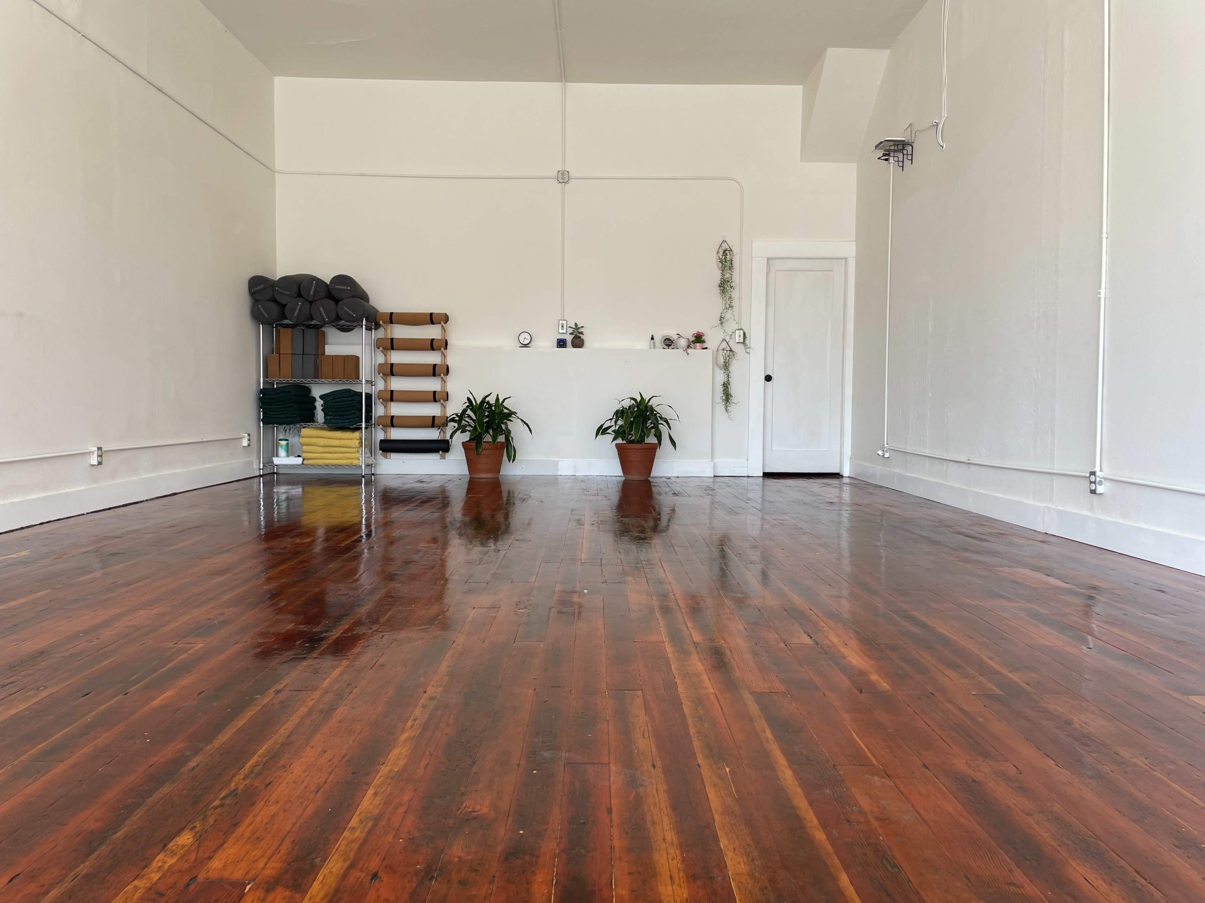 A spacious, empty room with polished wooden floors, two potted plants, and a wooden shelf against the wall.