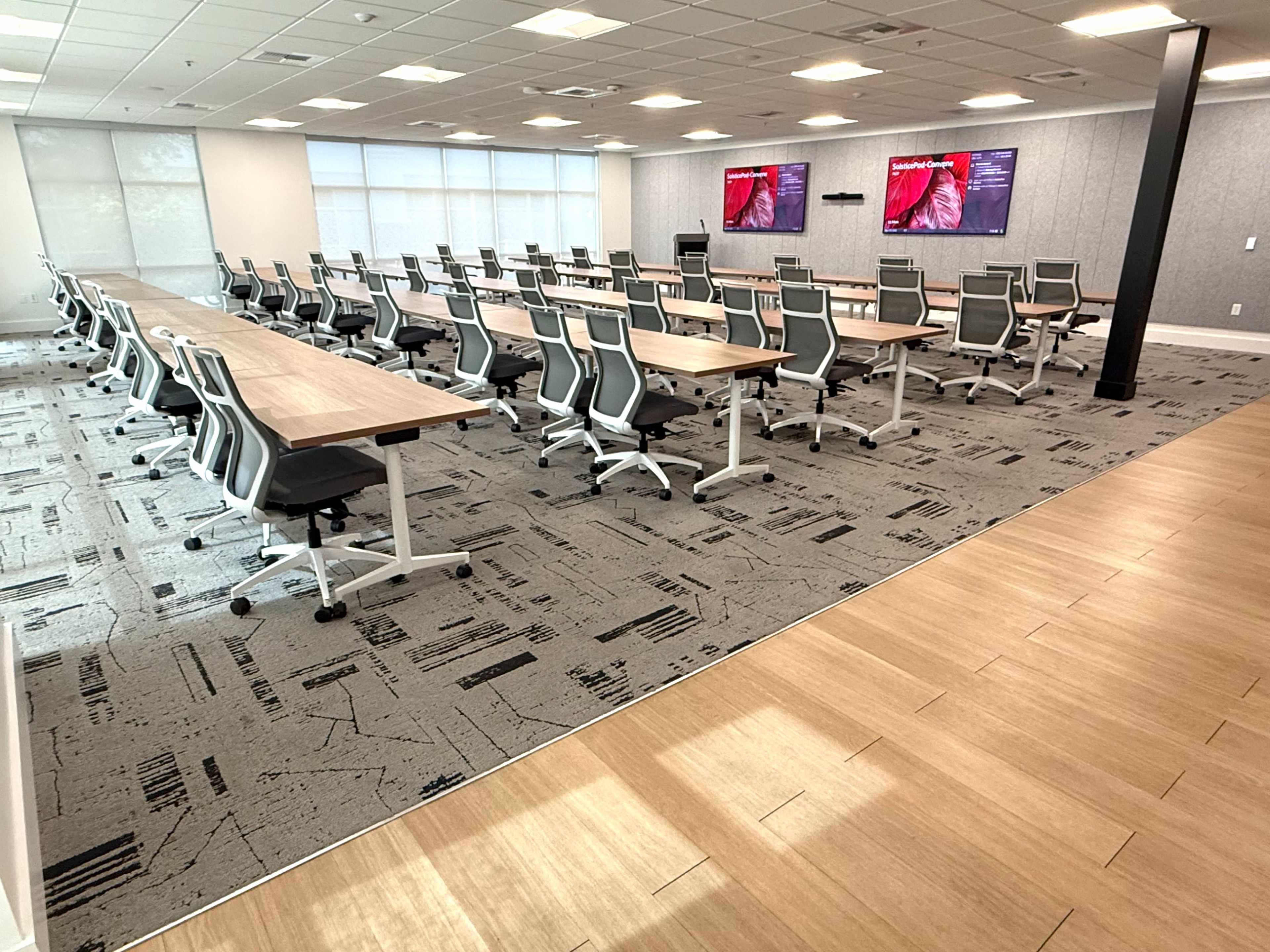 The image shows a modern conference room with rows of tables and chairs, equipped with two wall-mounted screens.