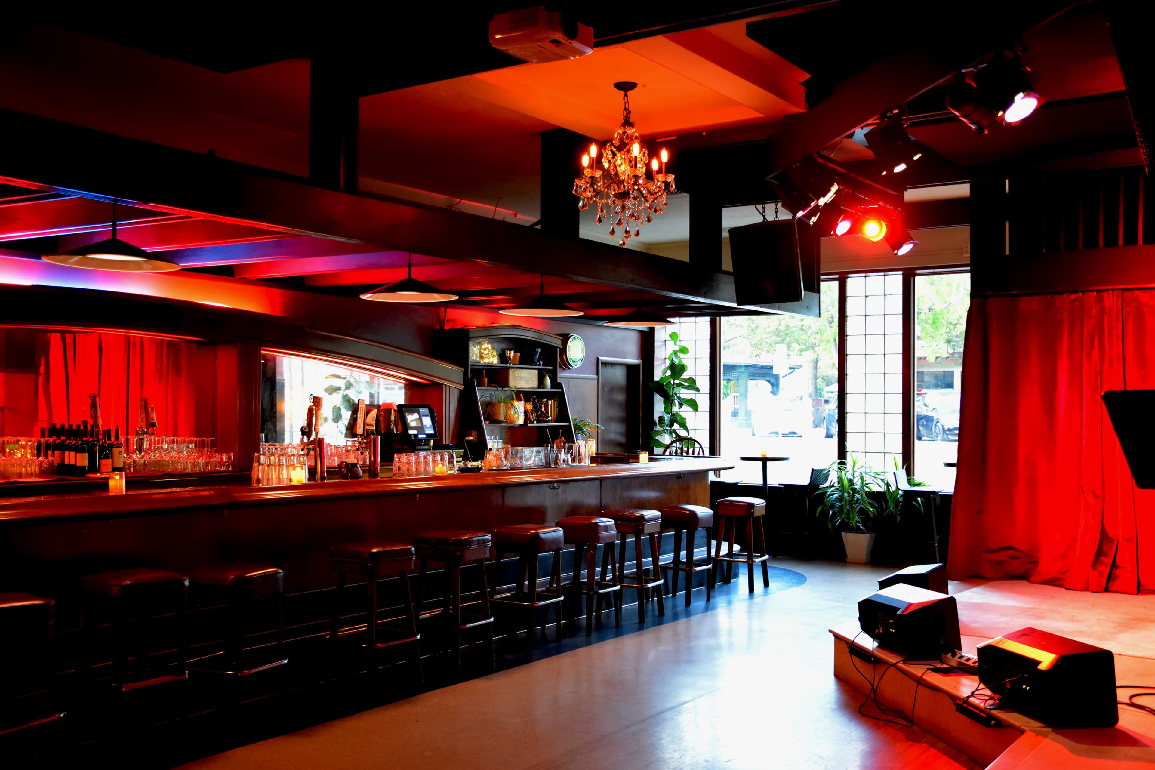 A stylish bar interior featuring a long counter with stools, a stage area with lights, and a red curtain.