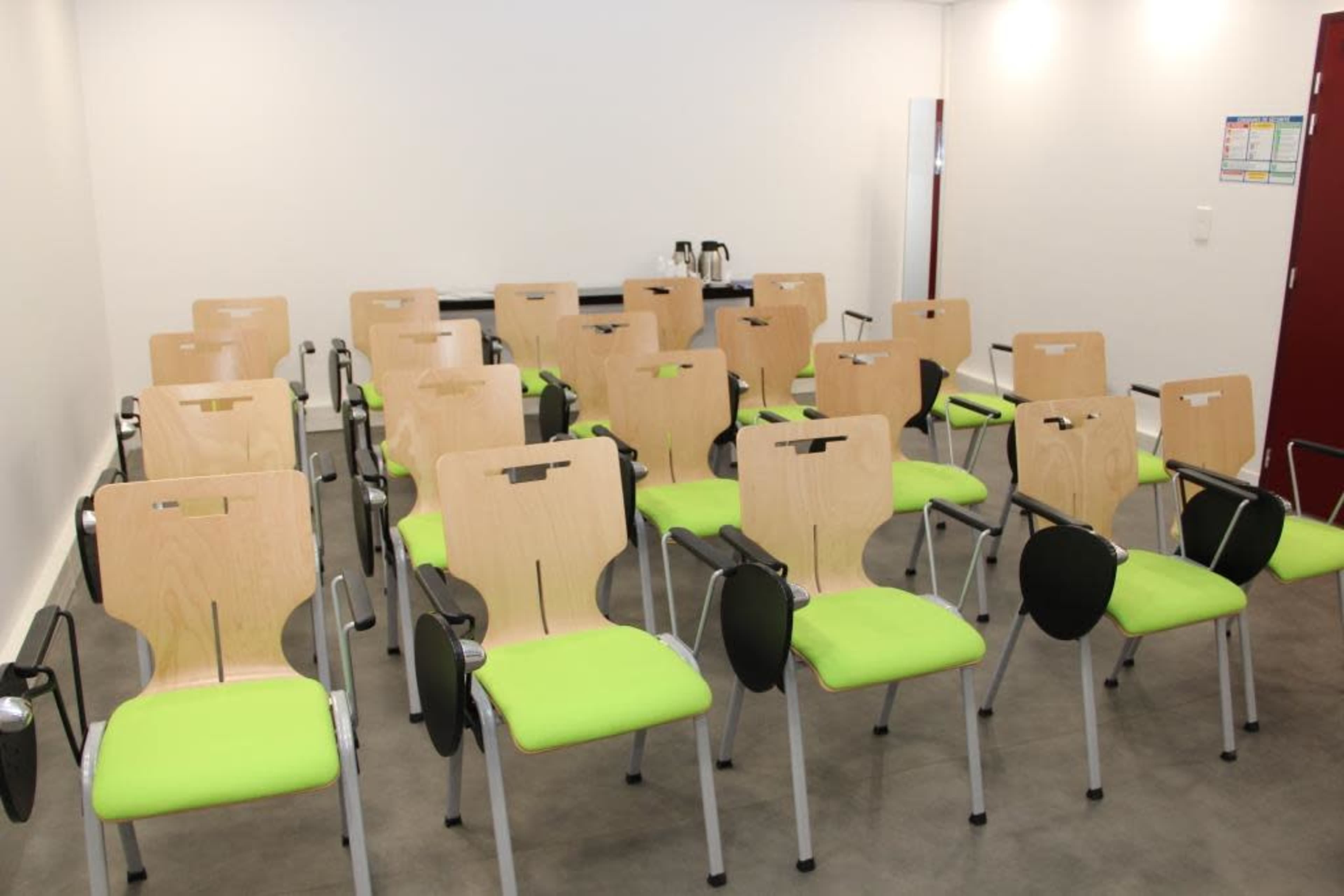 A row of empty chairs with wooden backs and green cushions is arranged neatly in a bright room.
