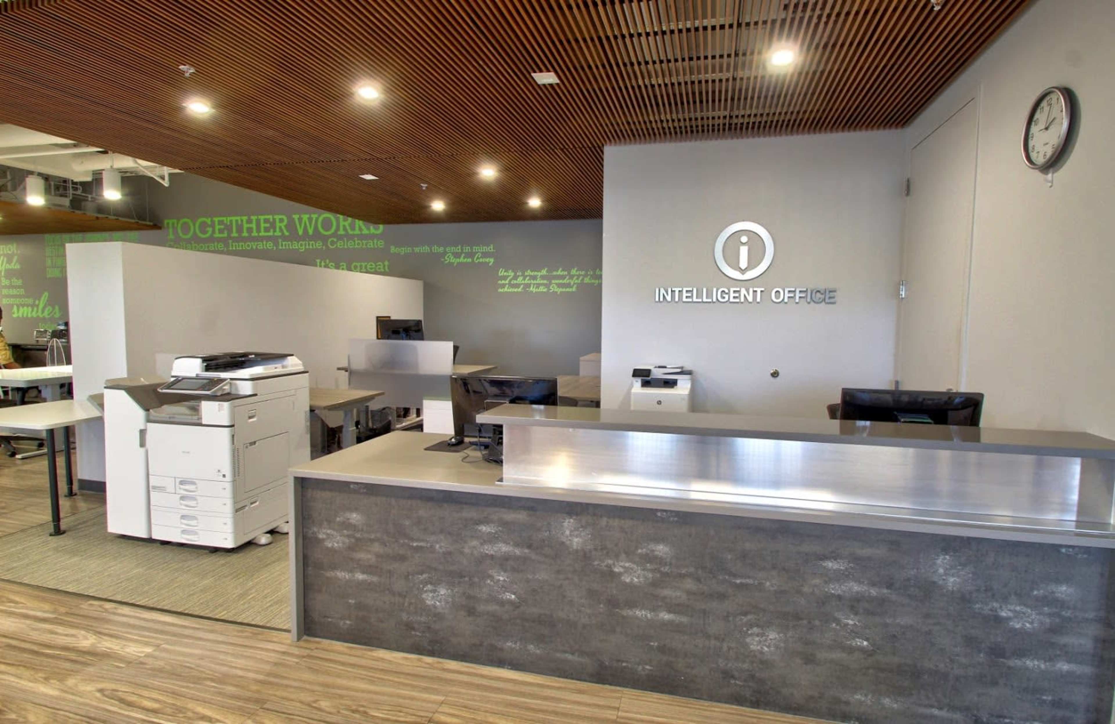 A modern office reception area with a sleek front desk, a printer station, and green wall graphics.