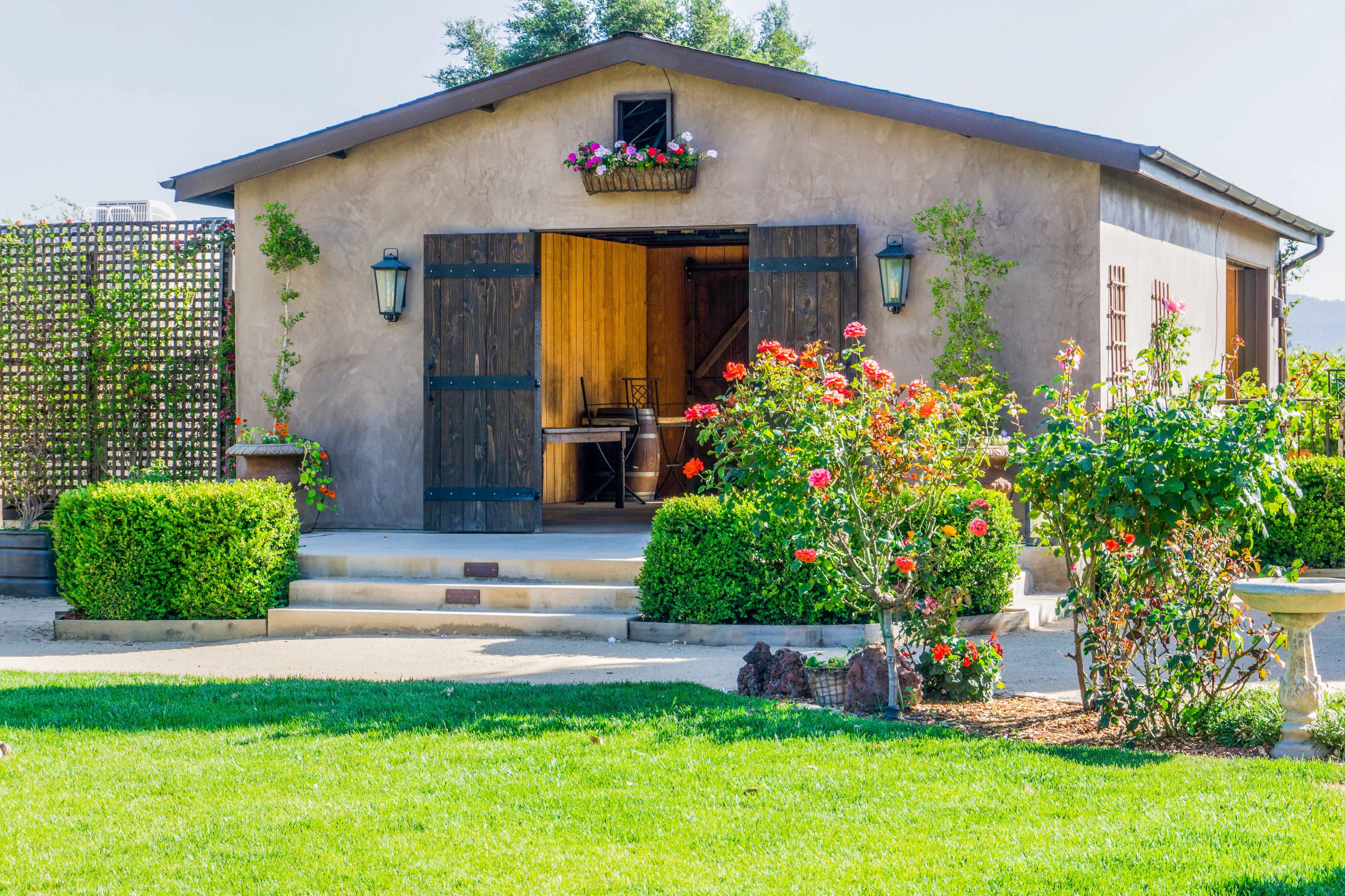 A rustic building with wooden doors adorned by flowers, surrounded by neatly trimmed greenery and a well-maintained lawn.