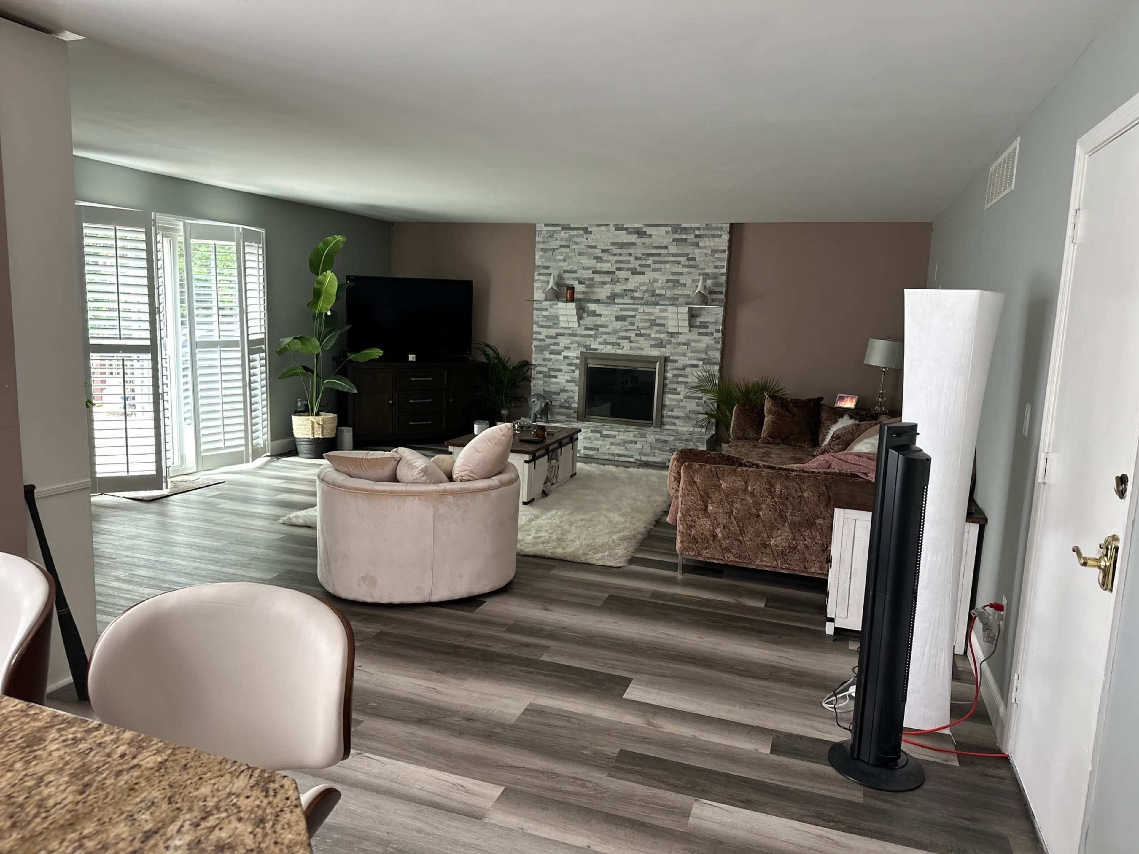 The image shows a living room with two seating areas, a stone fireplace, a television, and large windows with shutters.
