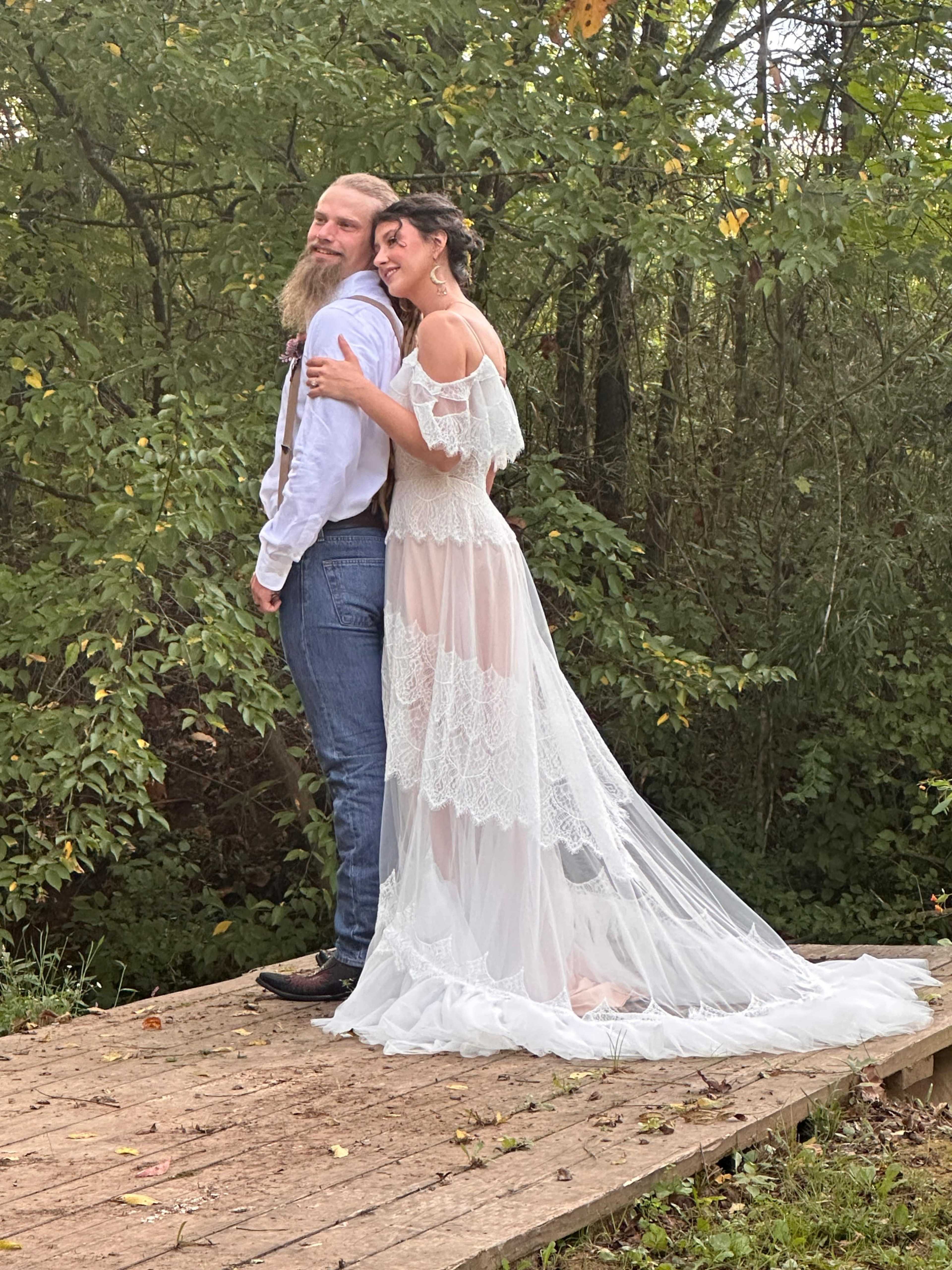 A couple stands back-to-back on a wooden platform surrounded by greenery, with the woman wearing a white lace gown and the man in a light shirt and suspenders.