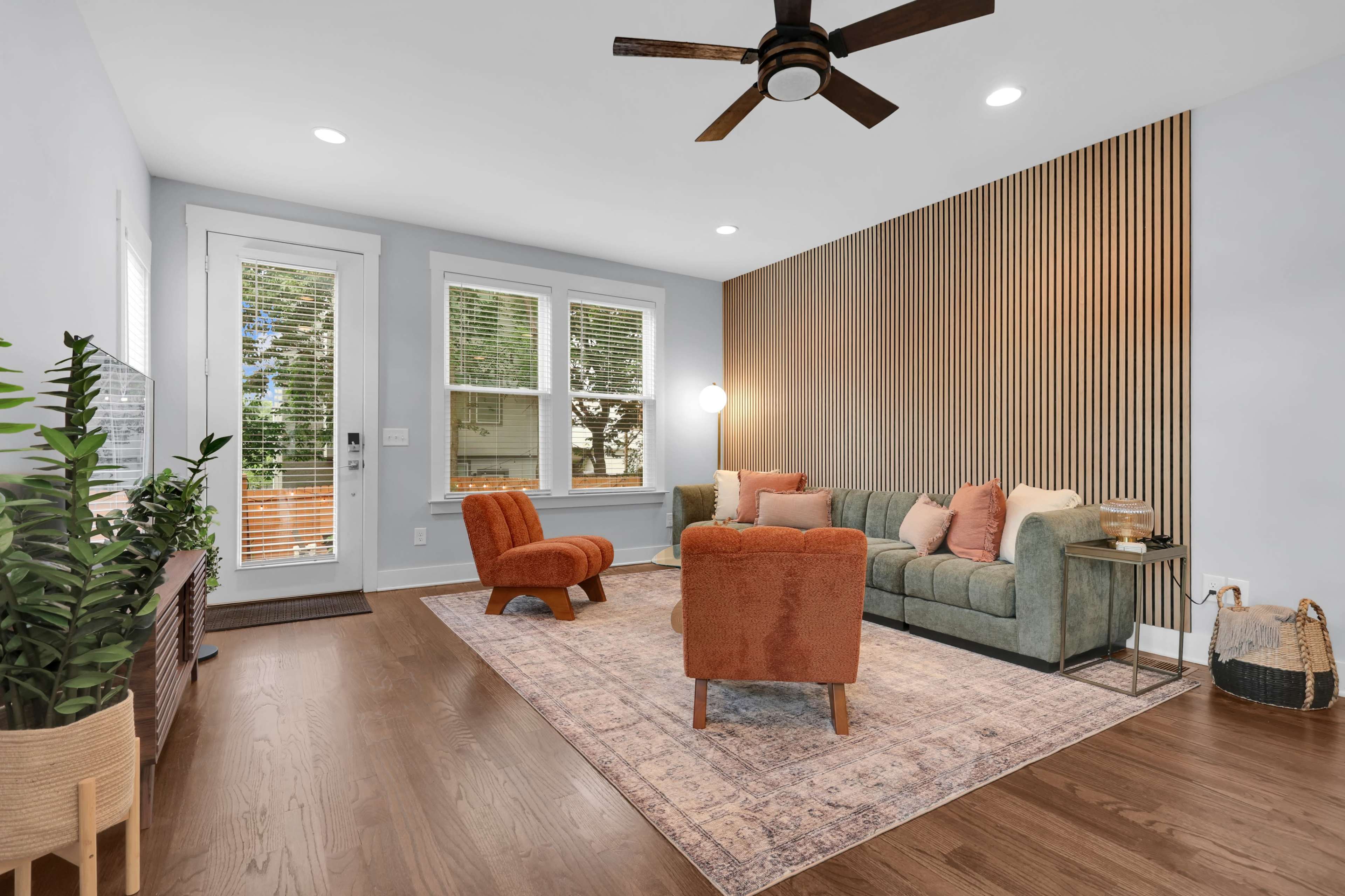 The image shows a modern living room featuring a green sofa, orange chairs, a wooden accent wall, and large windows that provide natural light.