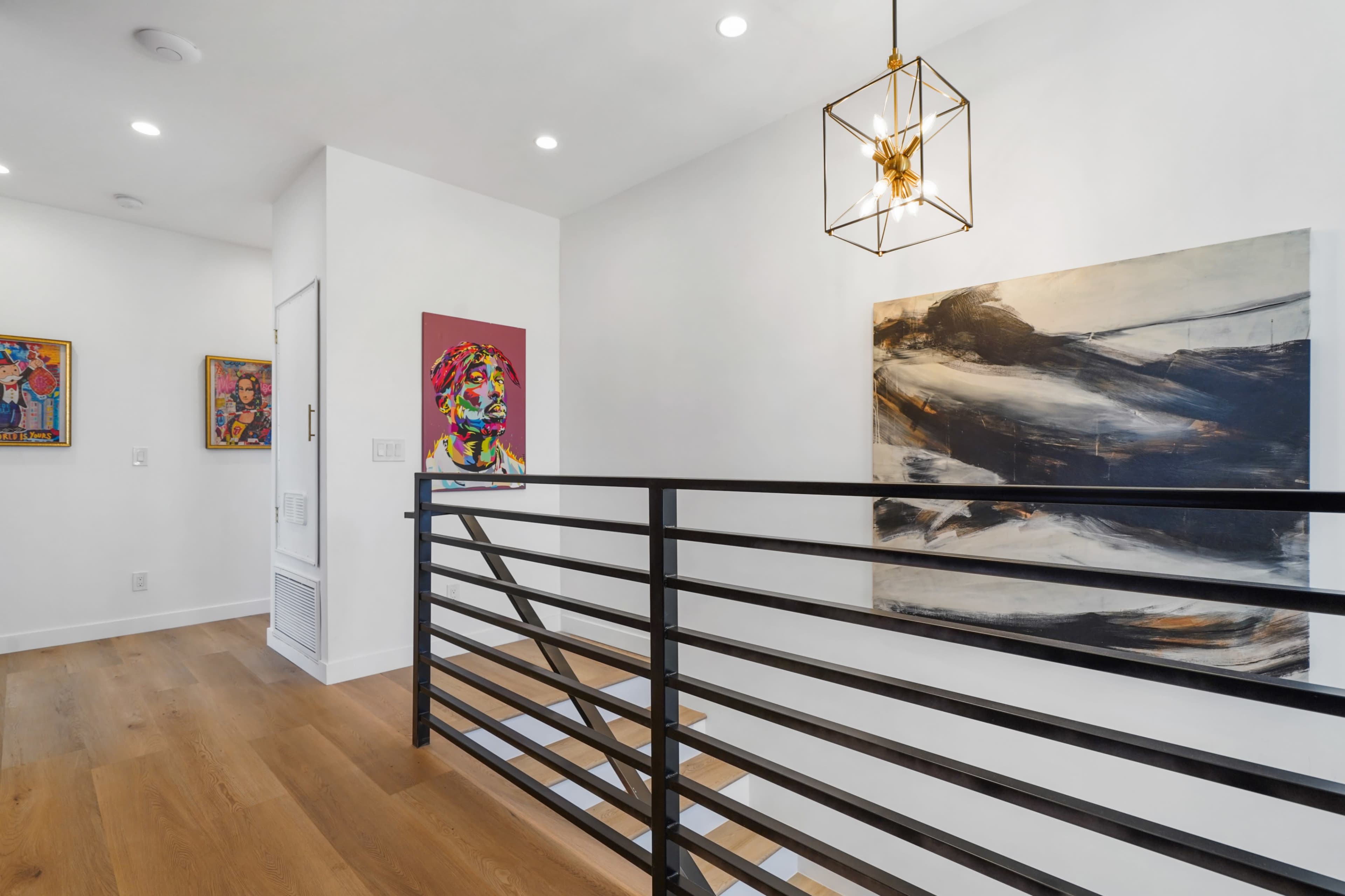 A well-lit hallway features a black metal railing, modern light fixture, and colorful artwork on white walls.