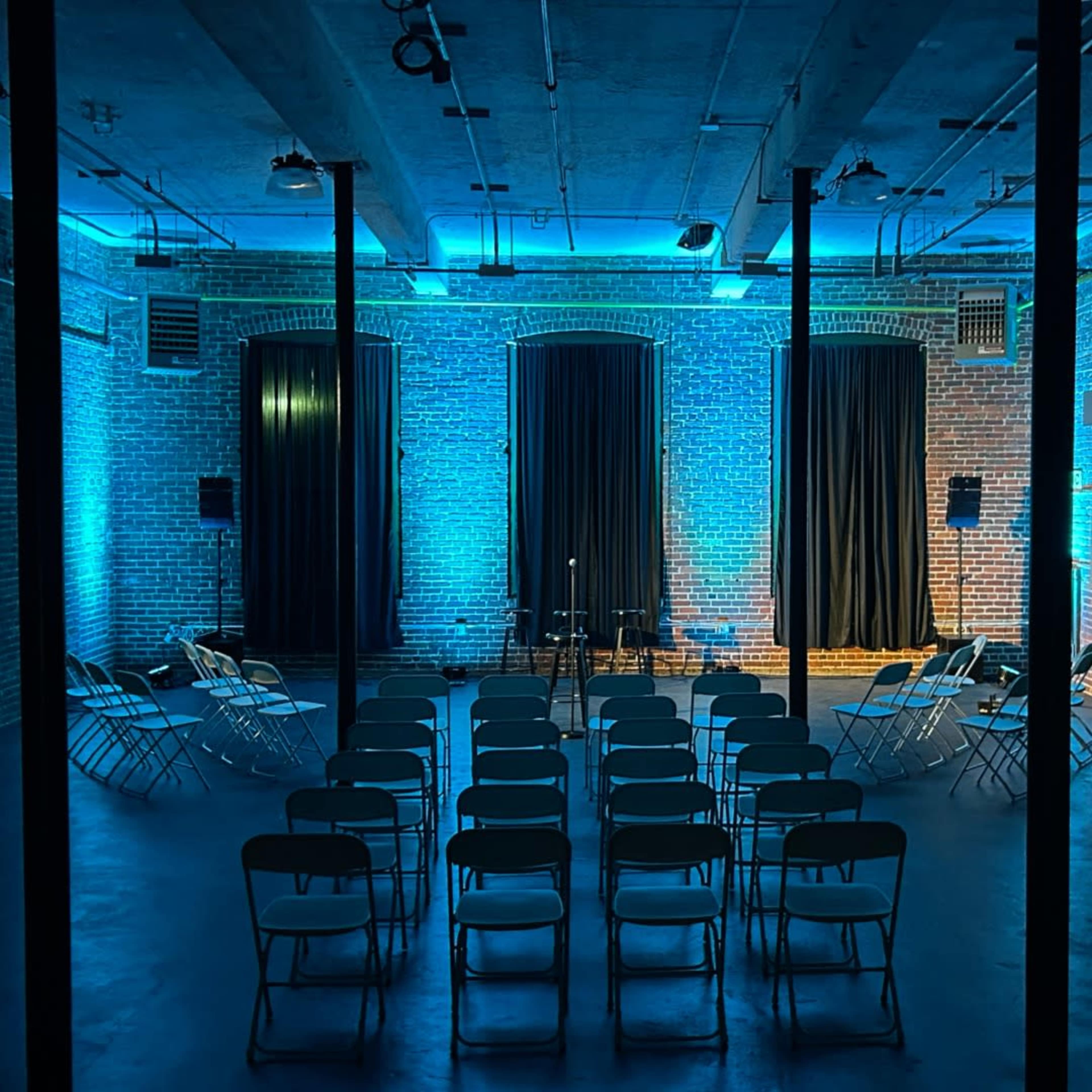 The image shows a dimly lit performance space with rows of metal chairs facing a stage, against a backdrop of exposed brick walls illuminated by blue lighting.