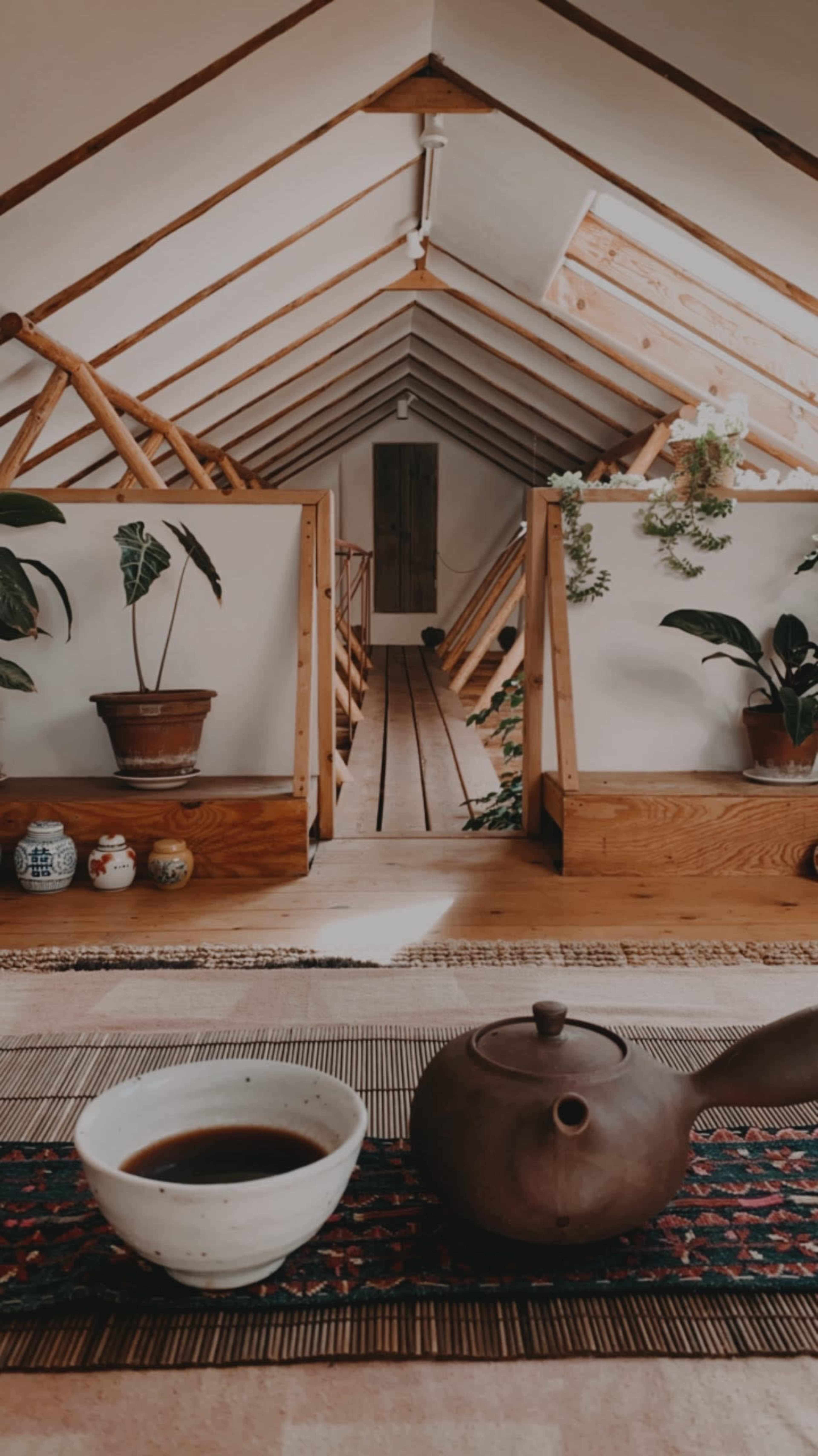 A wooden attic space with a central pathway, framed by plants and a tea set on a woven mat in the foreground.