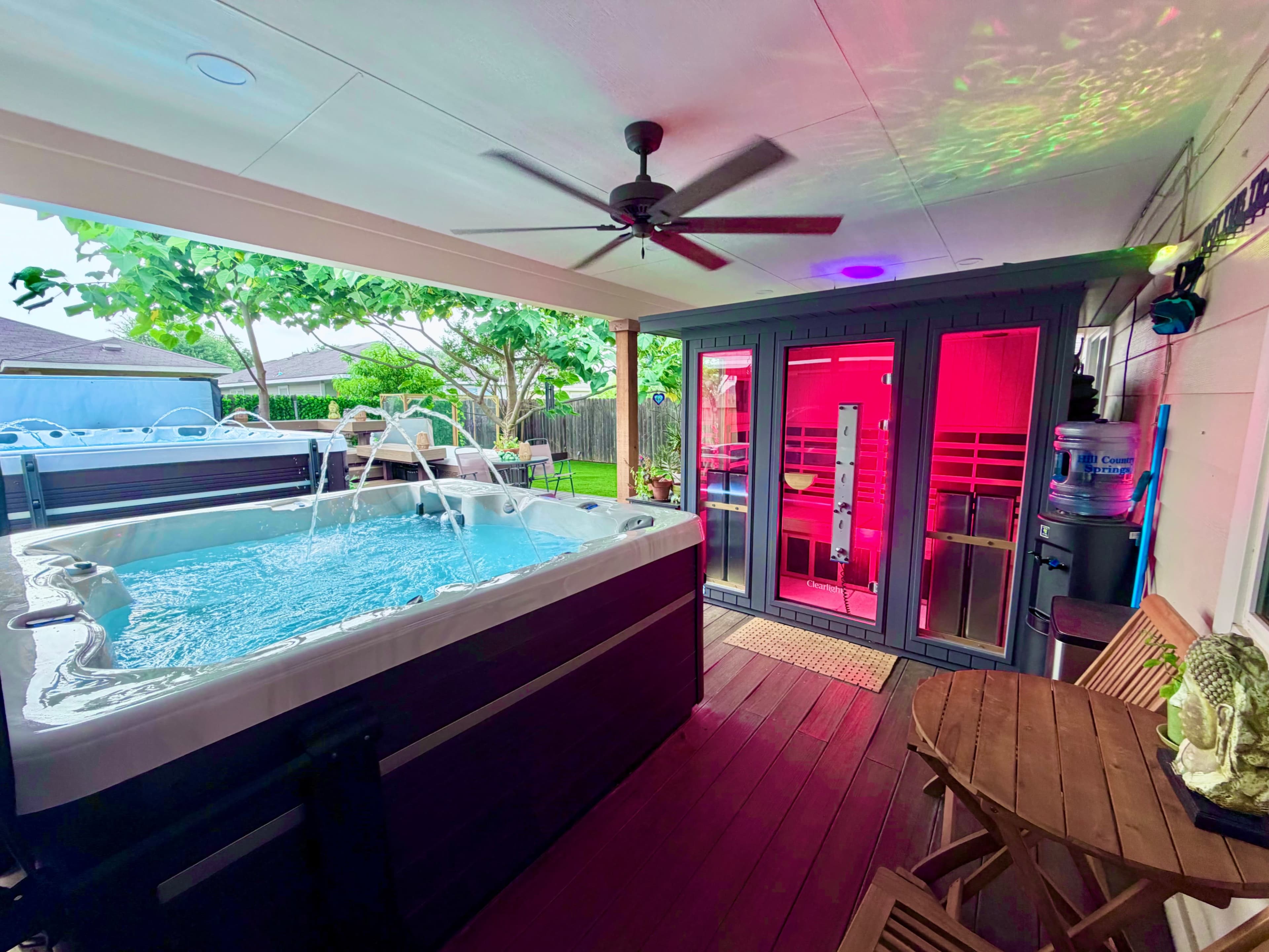 The image shows a backyard space featuring a hot tub with water jets, a sauna, and wooden seating under a covered area.