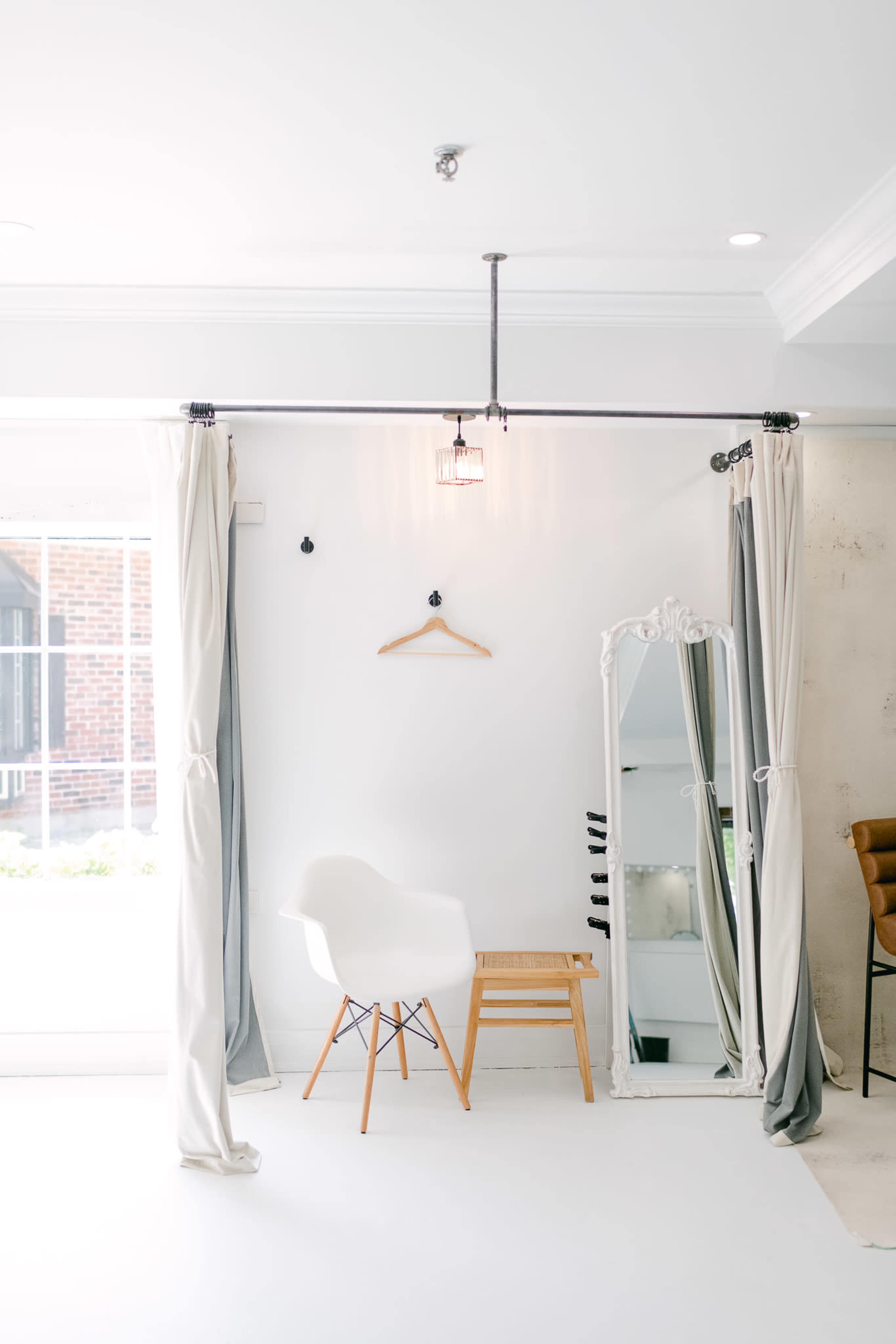 A minimalist interior design featuring a white chair, a wooden stool, a large mirror with a decorative frame, and curtains draped on either side of the space.