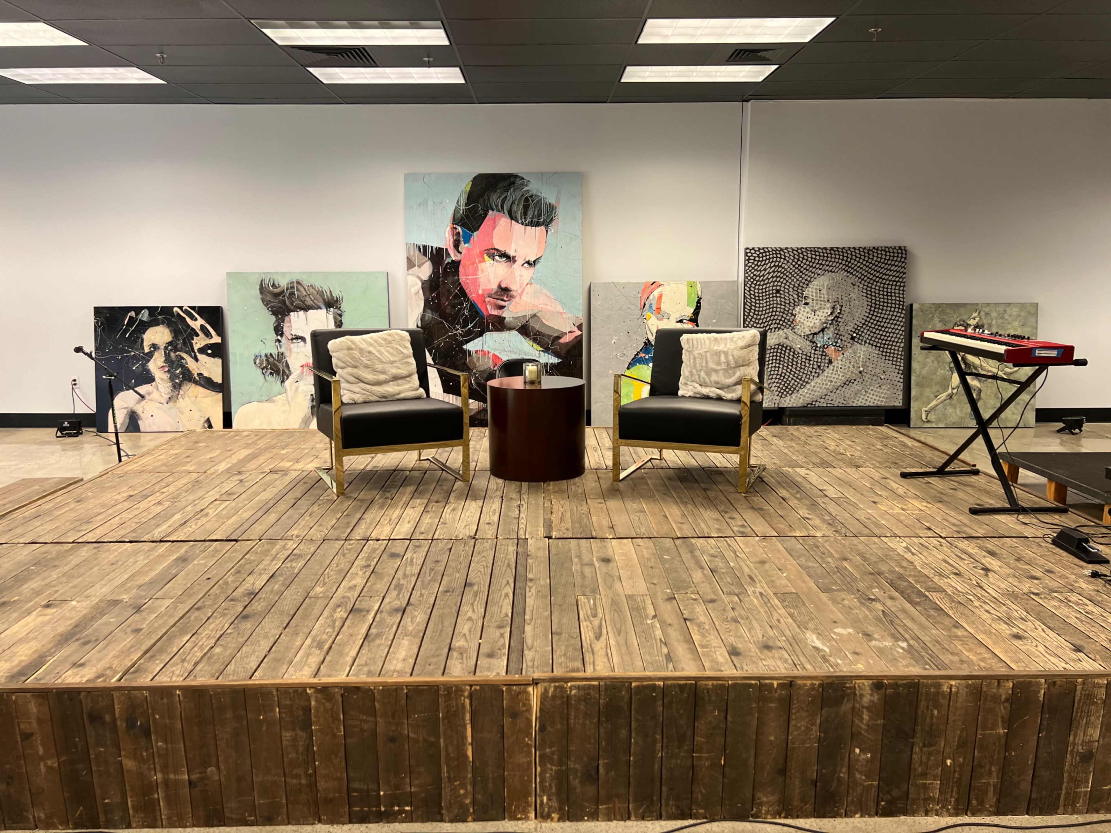 A stage with a wooden floor features two armchairs and a circular table, surrounded by multiple artistic portraits on the walls.