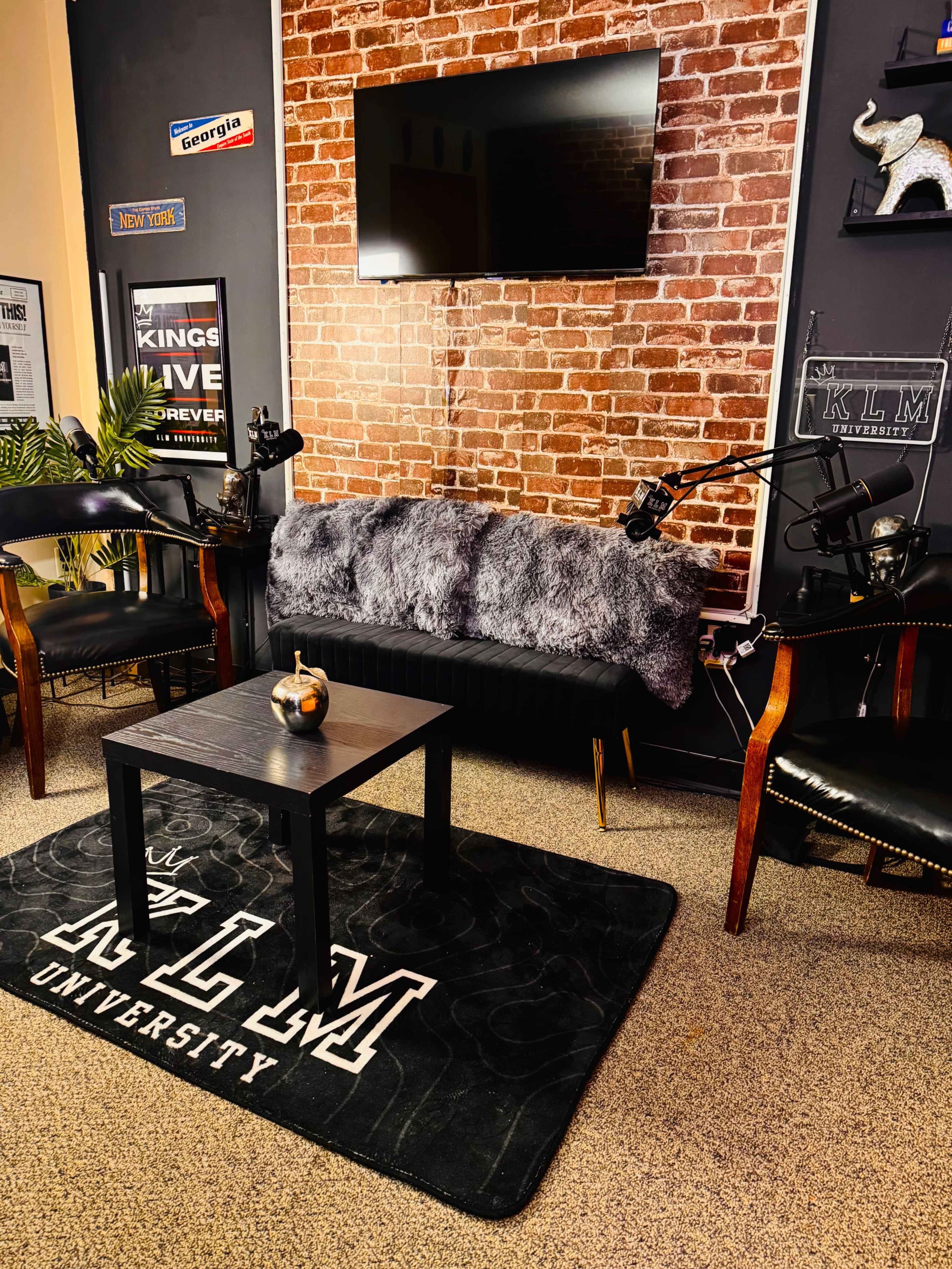 The image shows a cozy studio or podcast setup with a brick wall, a black table, four chairs, and two microphones positioned for recording.