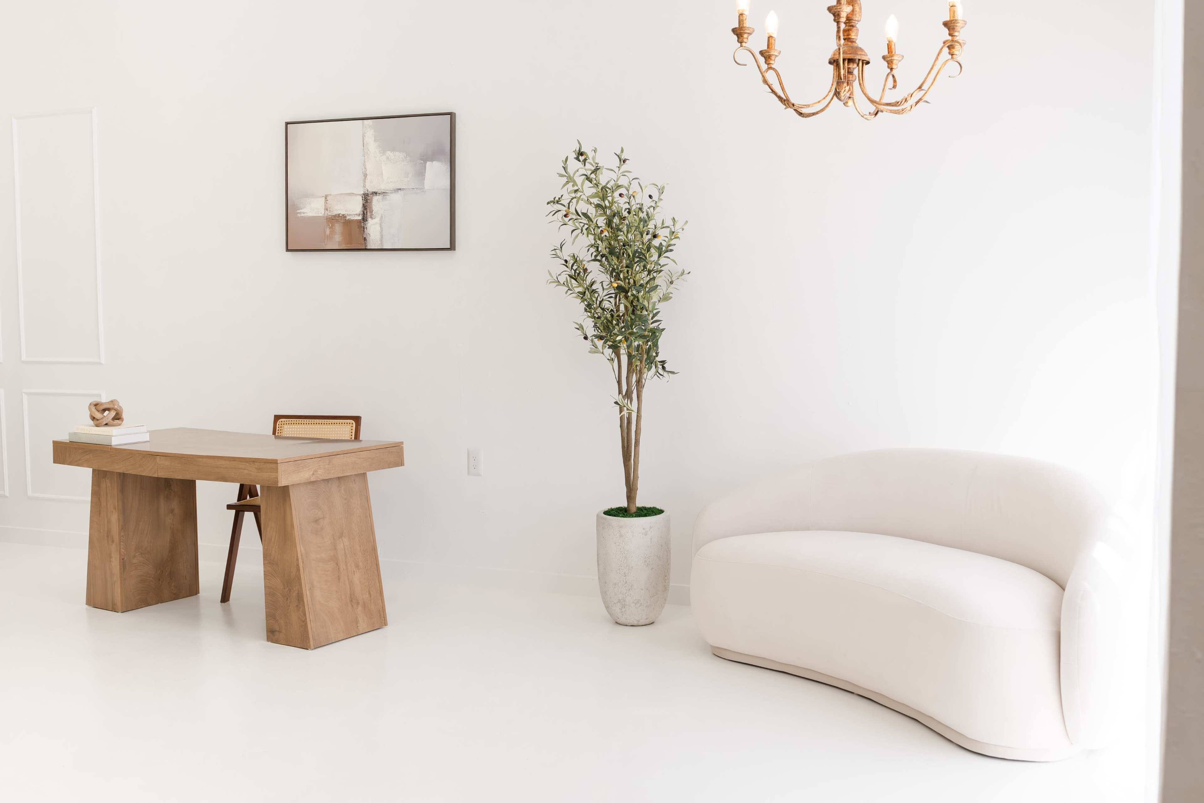 The image shows a minimalist office space featuring a wooden desk with a chair, a potted plant, and a curved white sofa, all set against a white backdrop.