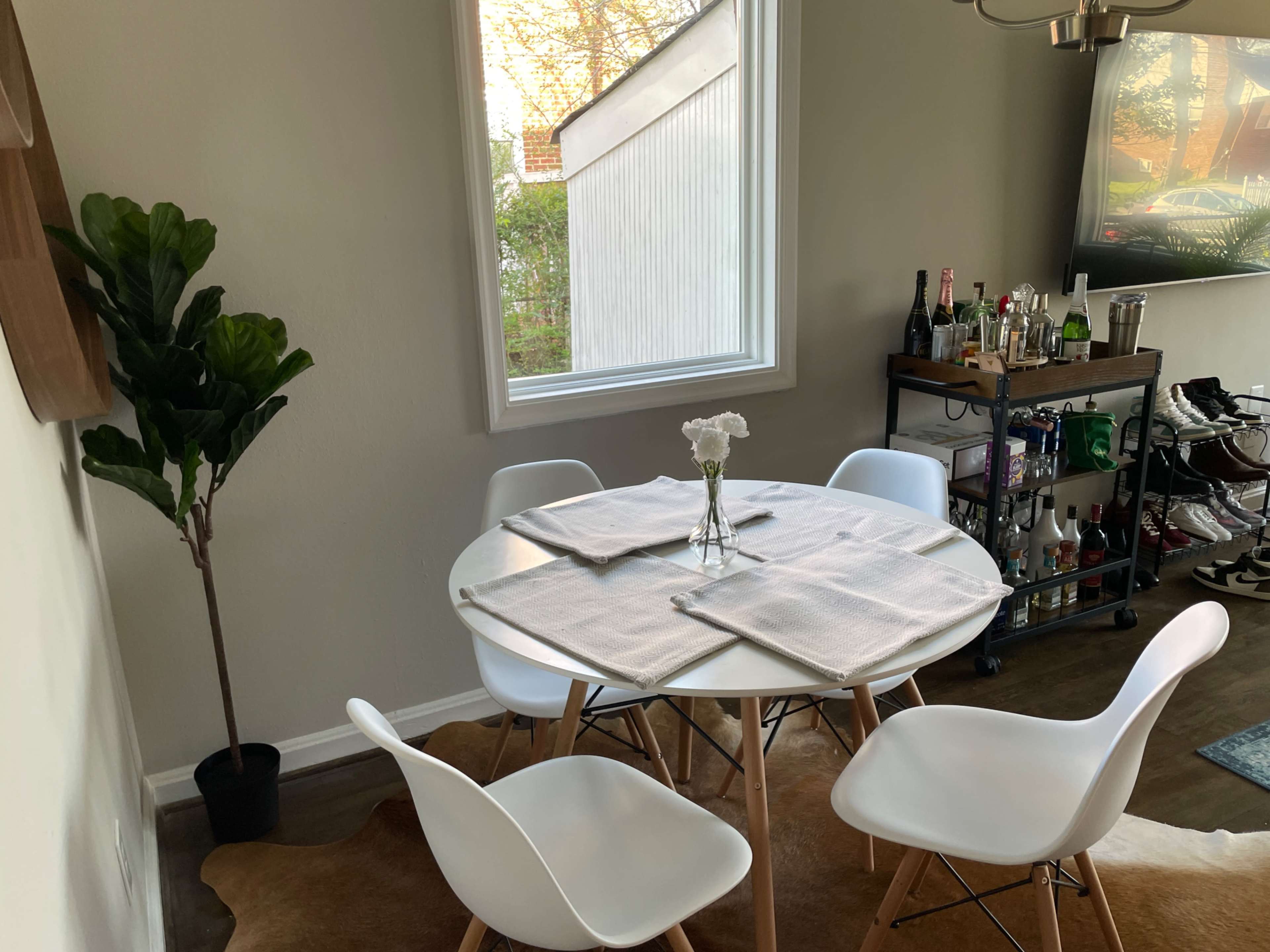A round dining table is set with placemats and a vase of flowers, surrounded by four white chairs and a large window that lets in natural light.