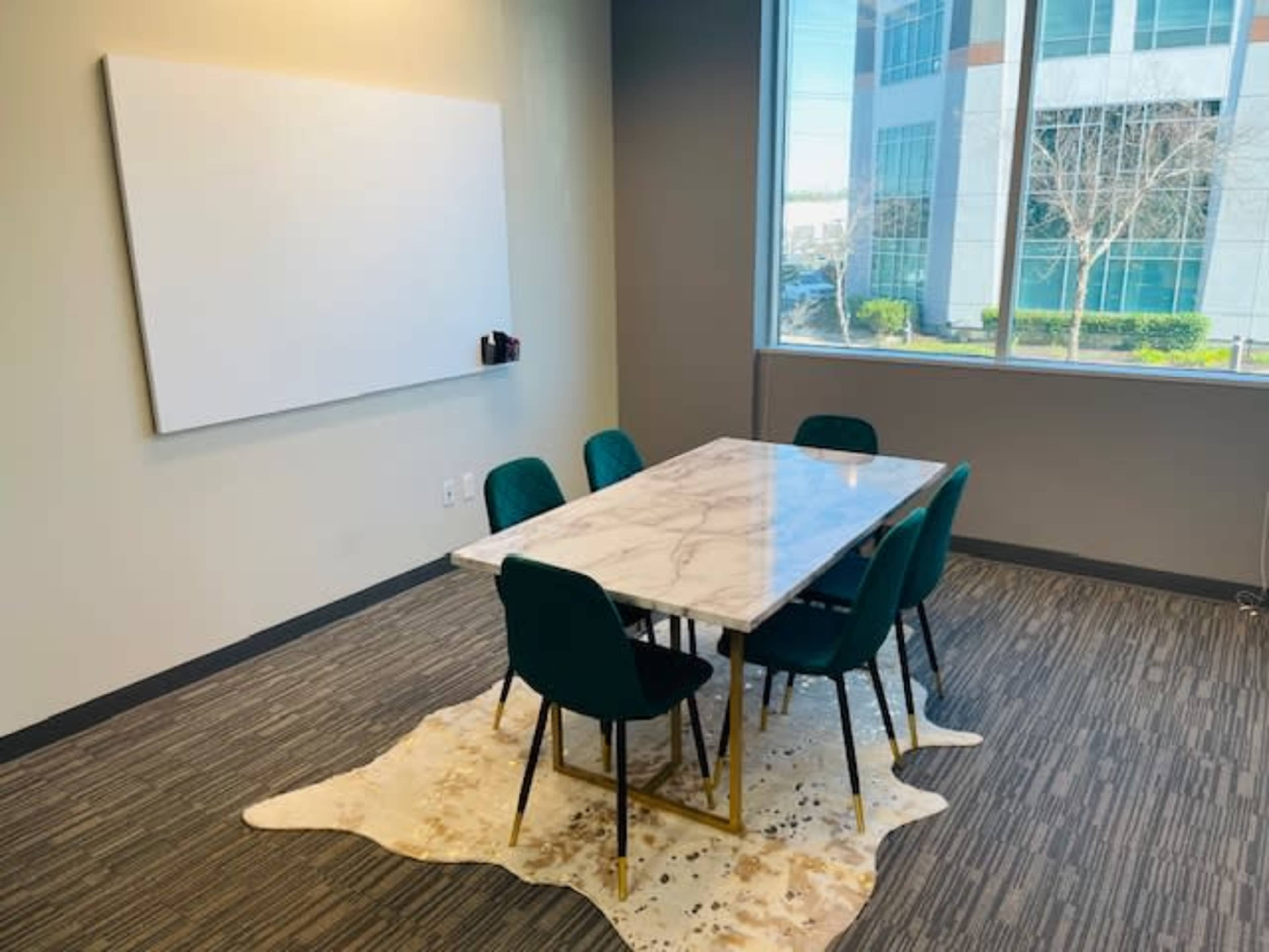 A modern meeting room features a marble-top table surrounded by green chairs, with a large whiteboard on one wall and a window overlooking an office building.