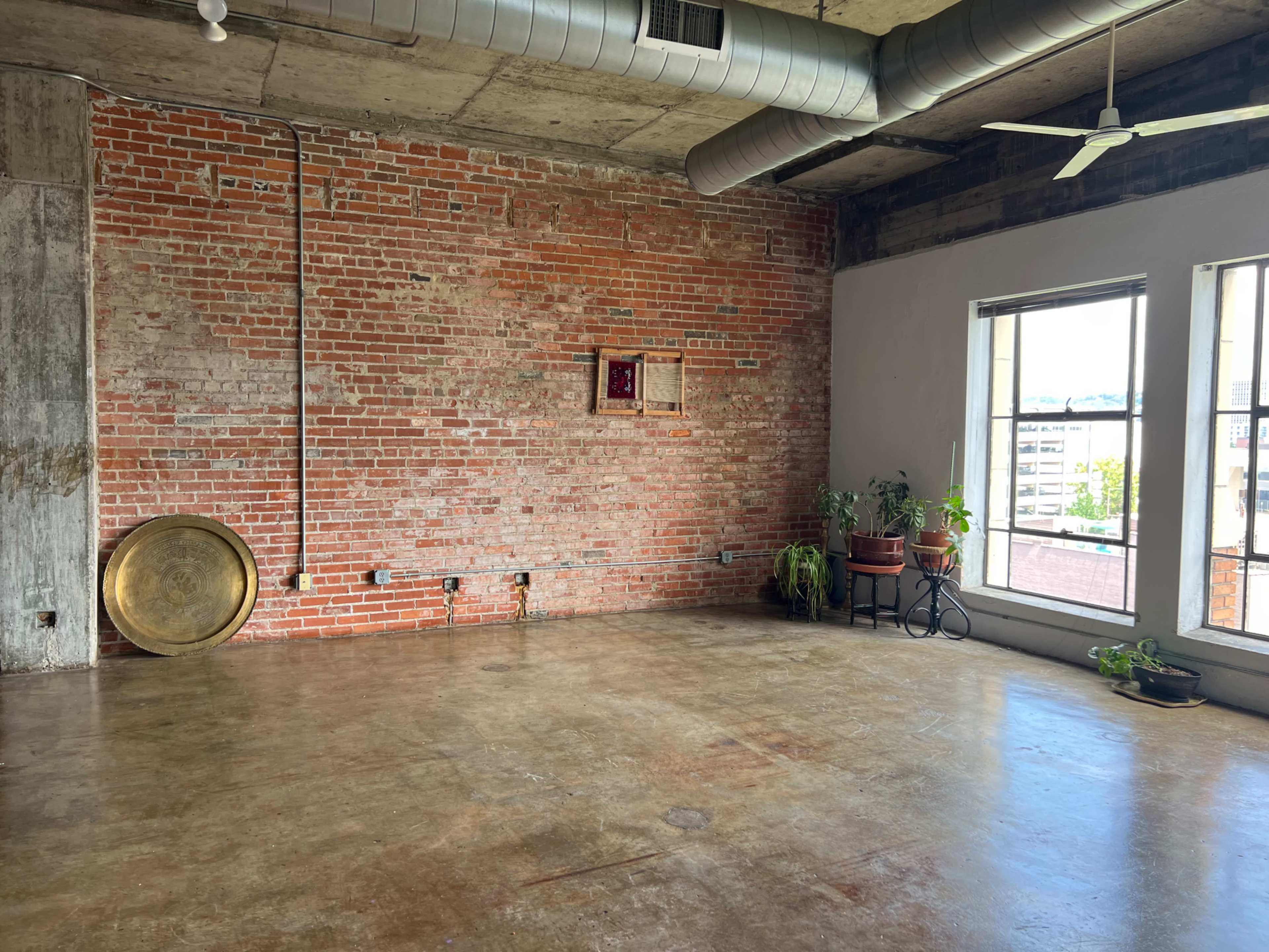 An industrial-style room featuring a brick wall, concrete flooring, and large windows with potted plants nearby.