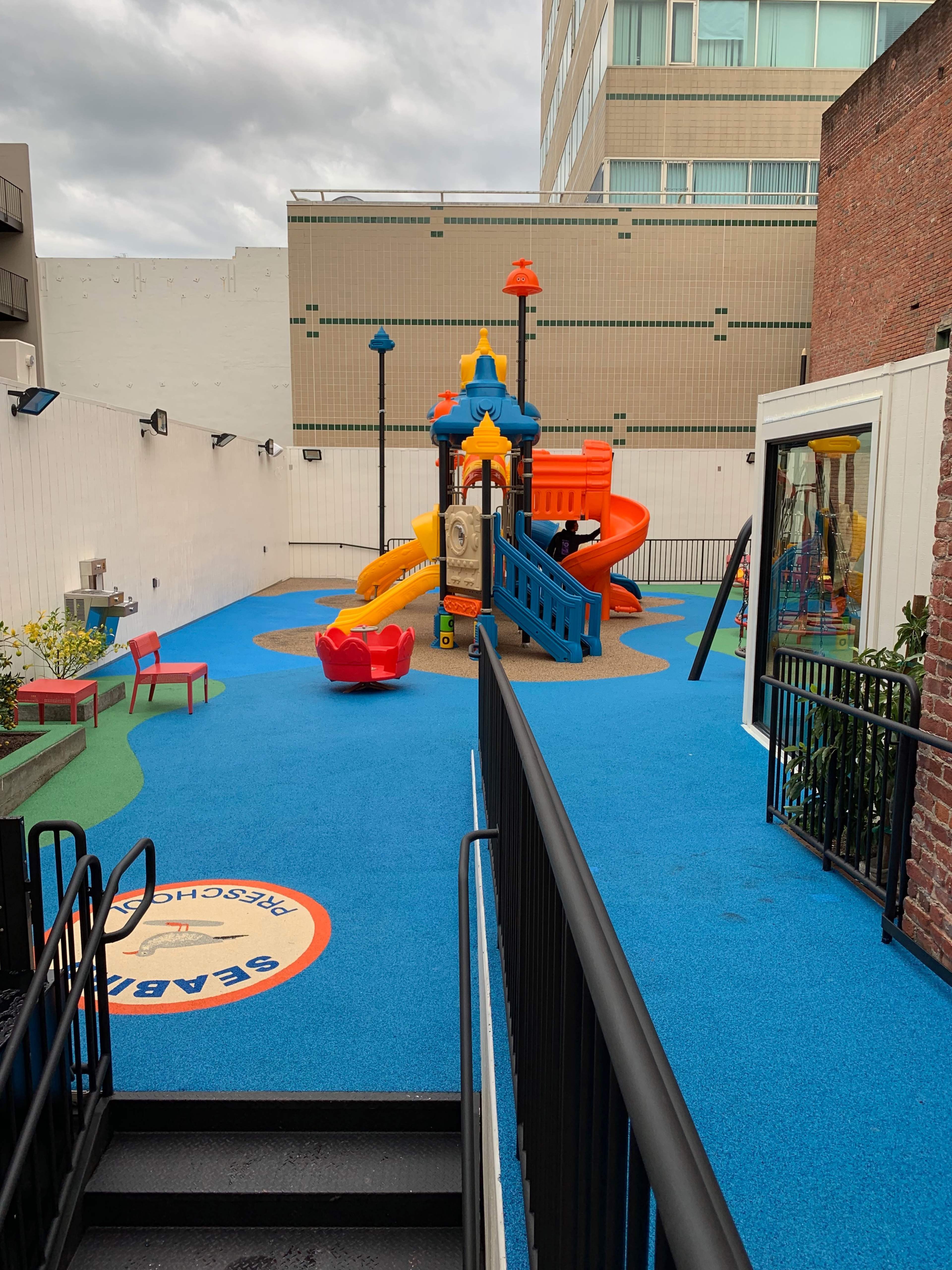 A colorful playground with a slide and climbing structure set on a blue rubber surface, surrounded by a low wall and nearby buildings.