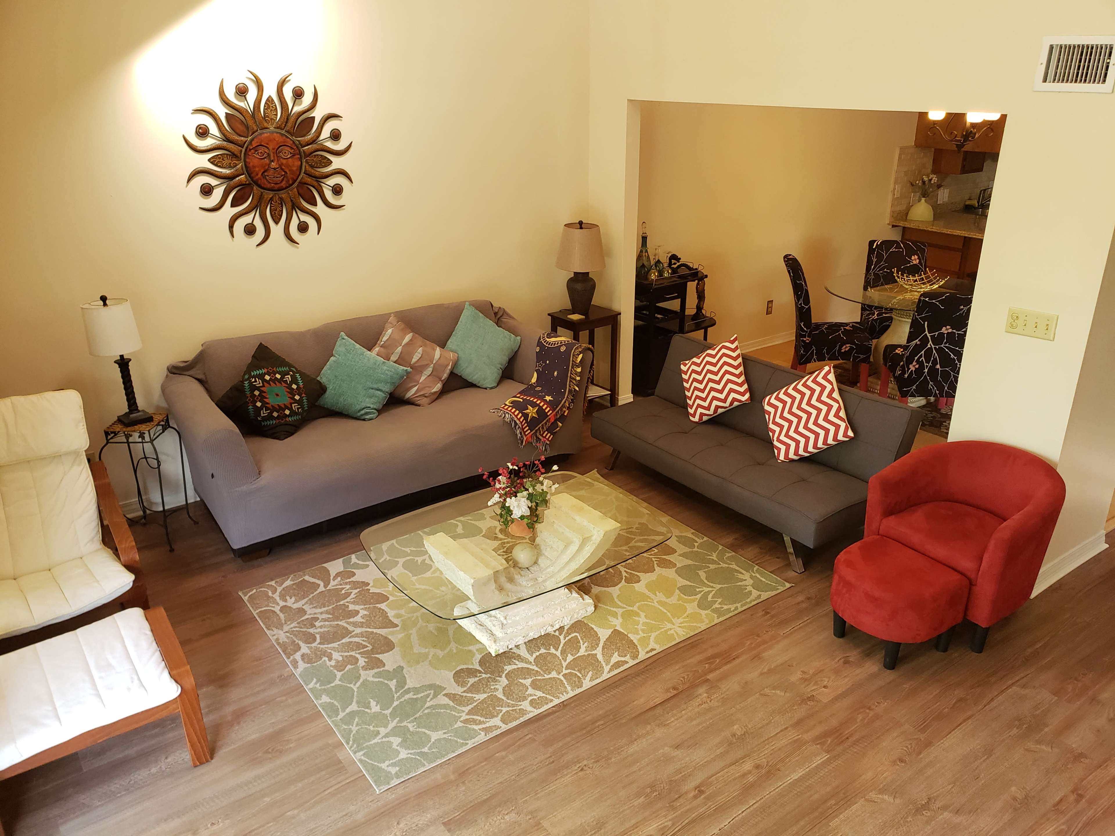 The image shows a living room area with two sofas, a glass coffee table, a decorative rug, and a wall-mounted sun sculpture.