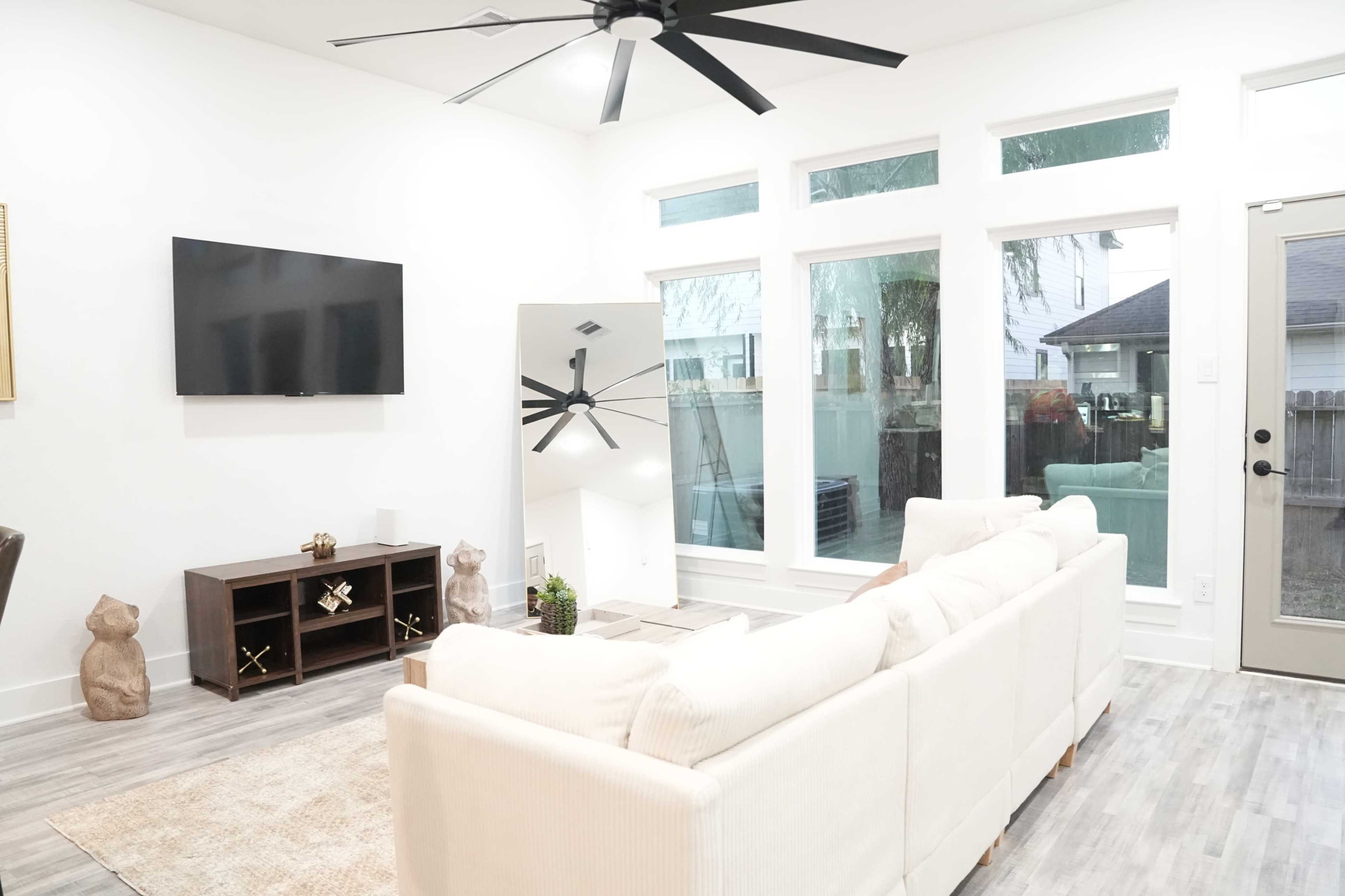 The image shows a modern living room with a large white sofa, a flat-screen TV mounted on the wall, and large windows allowing natural light to enter.
