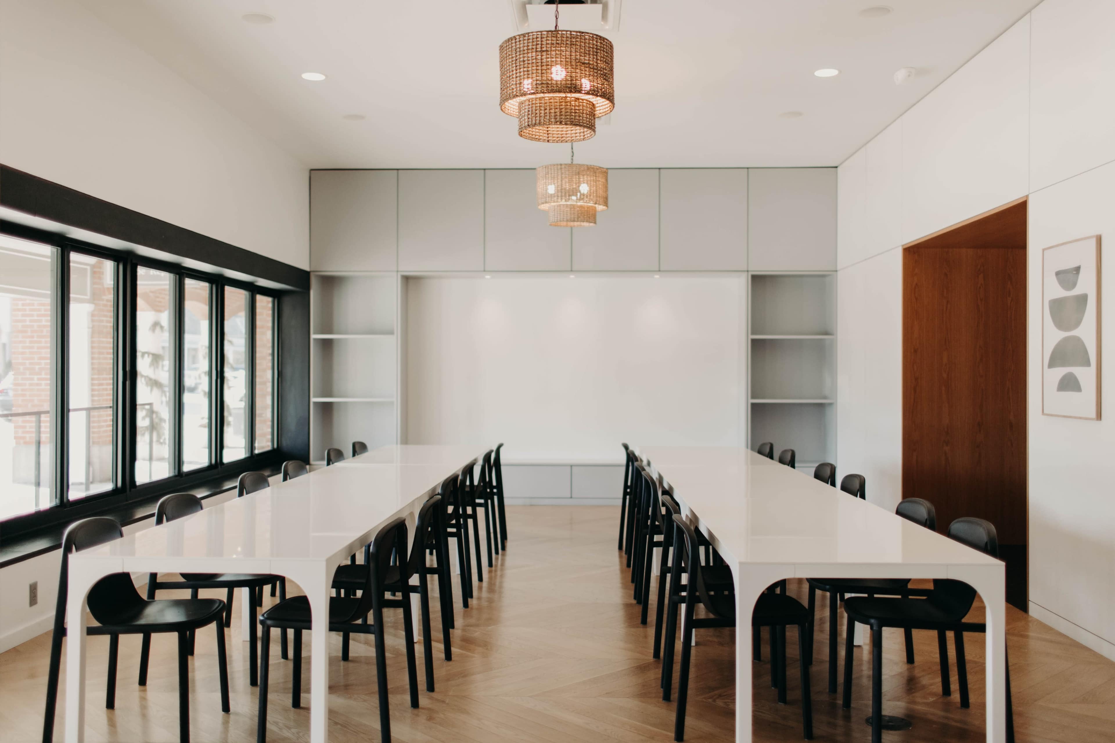 A modern conference room features a long white table surrounded by black chairs, illuminated by stylish pendant lights.