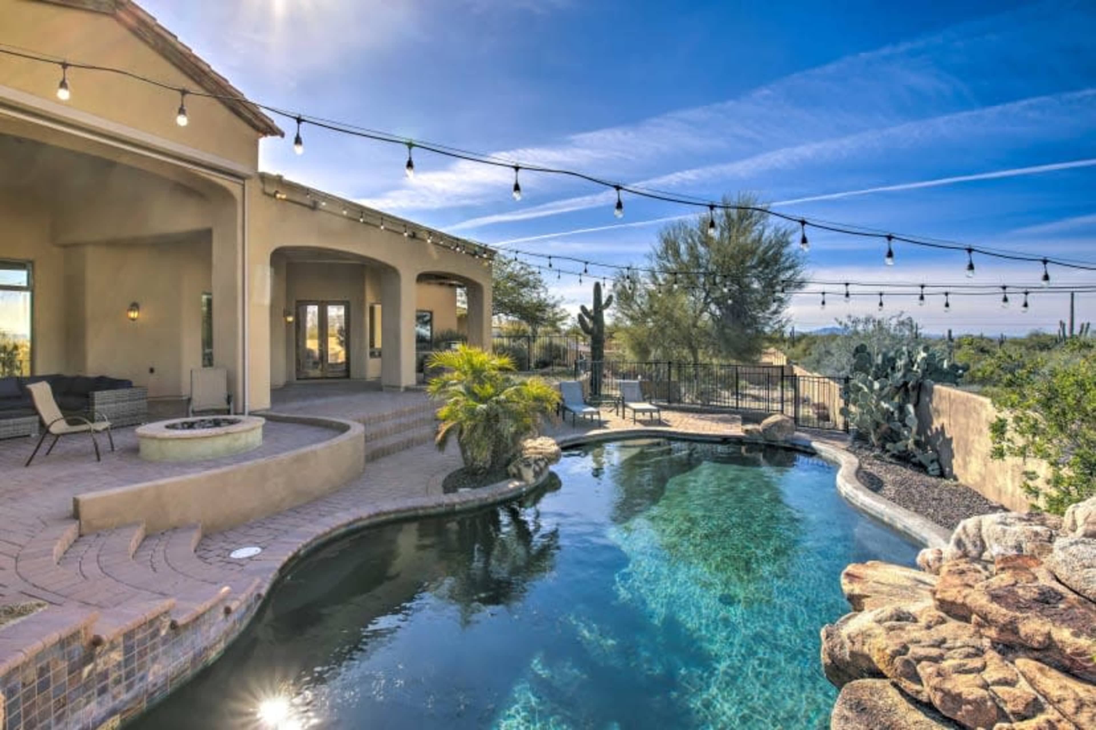 A backyard pool area with a hot tub, surrounded by desert landscaping and strung lights under a clear blue sky.