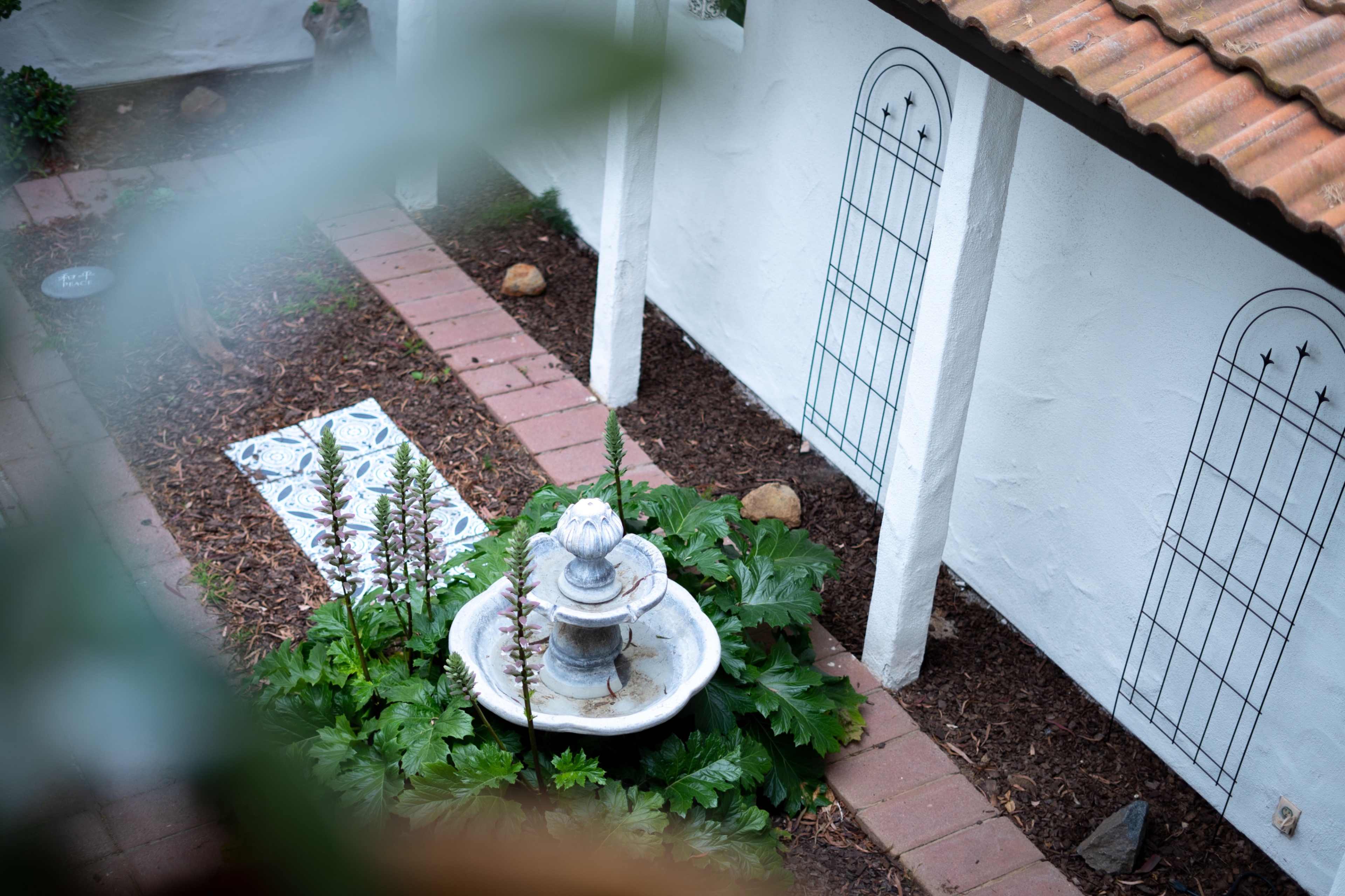 A stone fountain surrounded by greenery is positioned in a garden with brick pathways and white walls.