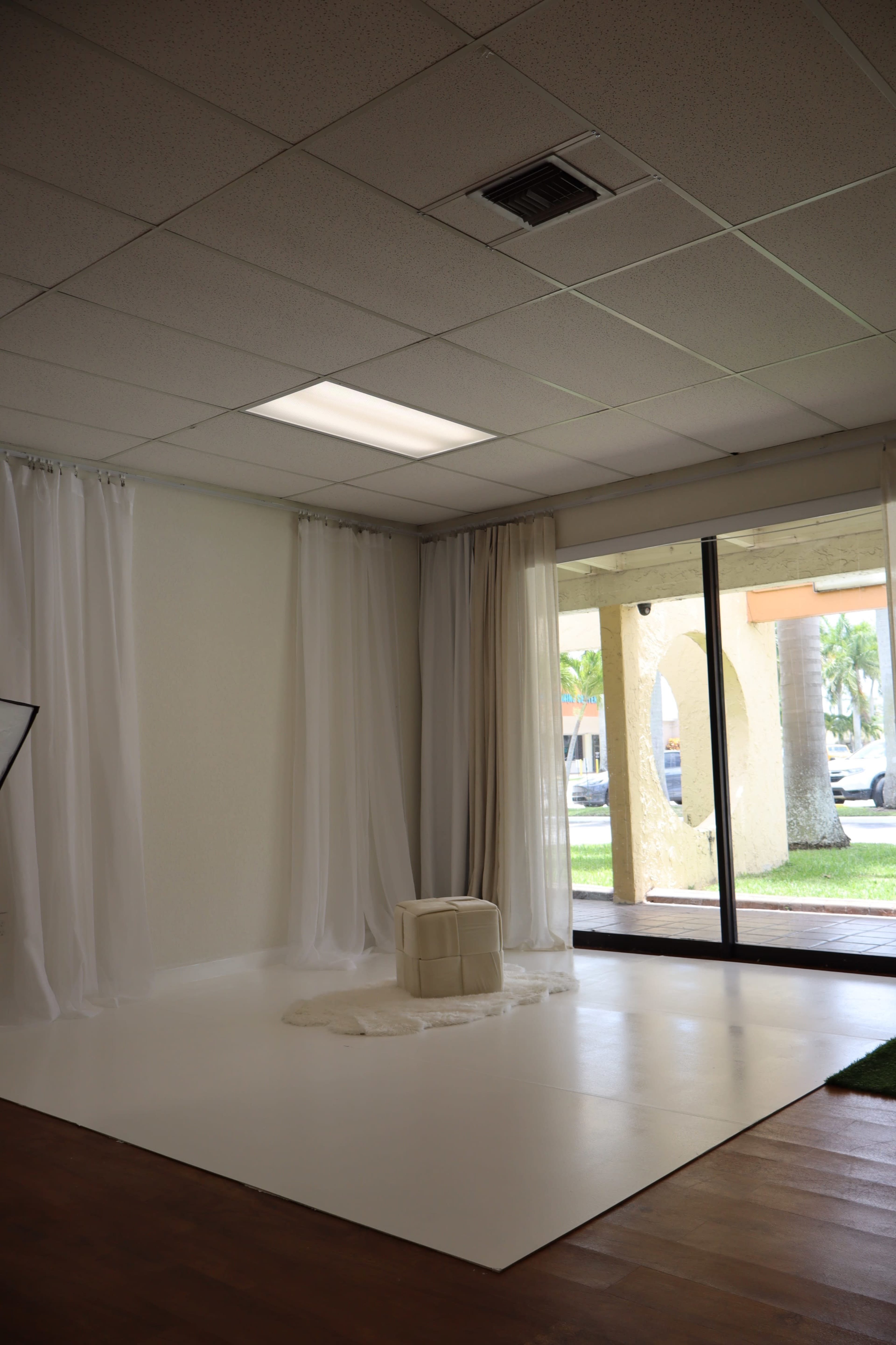 The image shows a spacious room with white walls, sheer curtains, a white floor, and a small stool in the center, illuminated by natural light coming through large windows.