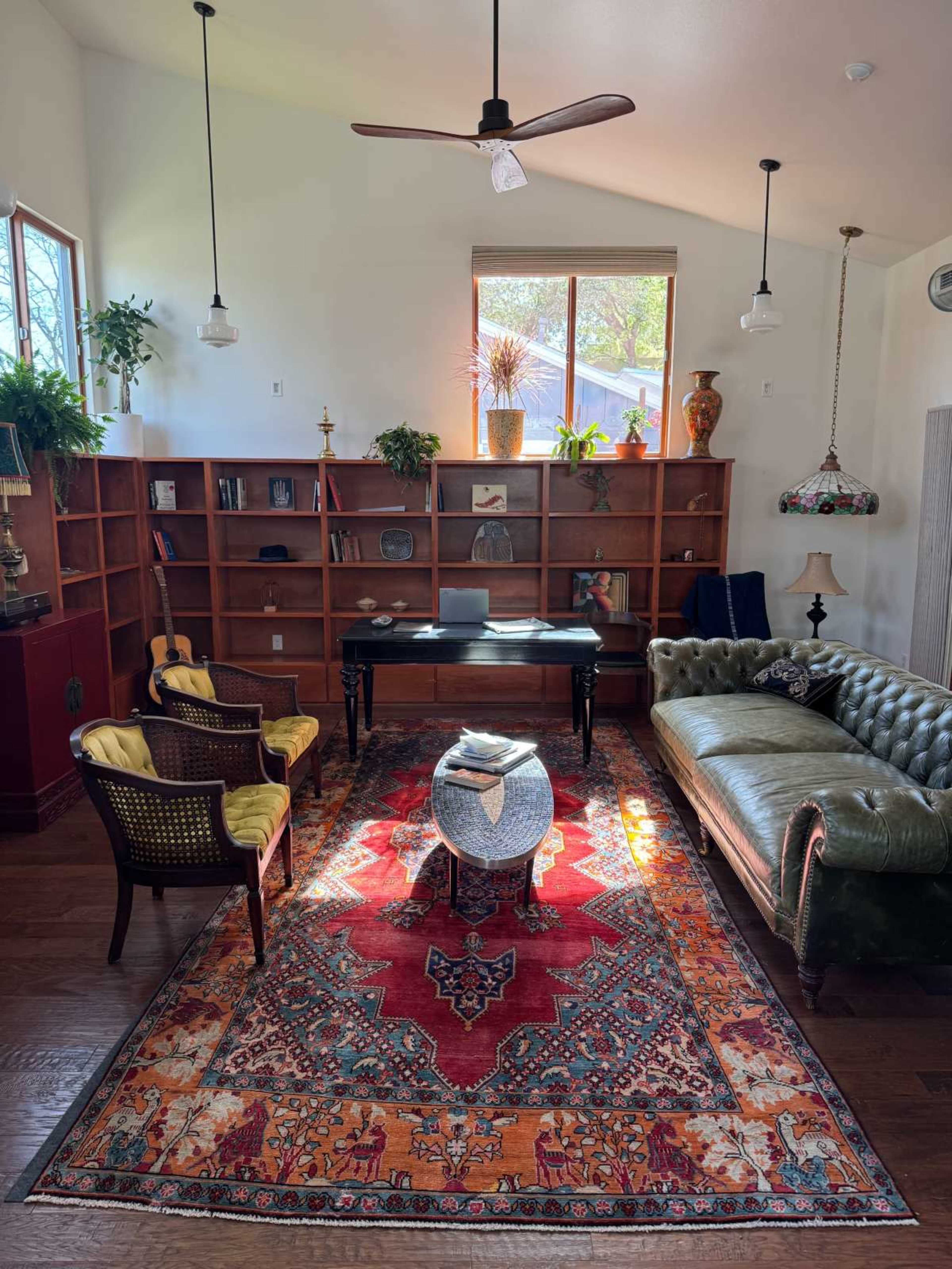 The image shows a well-lit room with a green leather sofa, two patterned chairs, a black table, and a large area rug, all arranged around a wooden bookshelf.