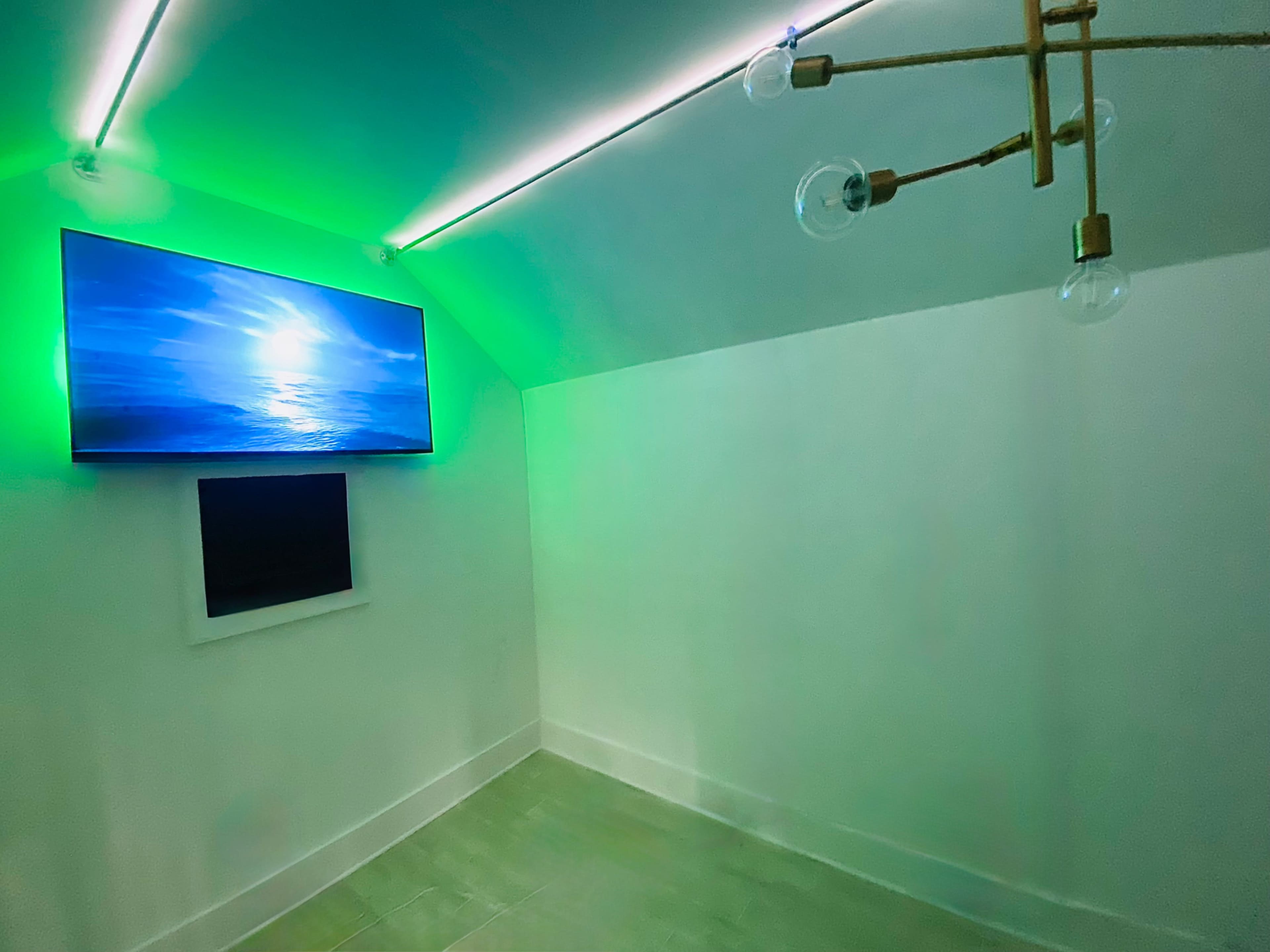 The image shows a minimalist room with a television displaying a seascape and a modern light fixture, both illuminated by green lighting.