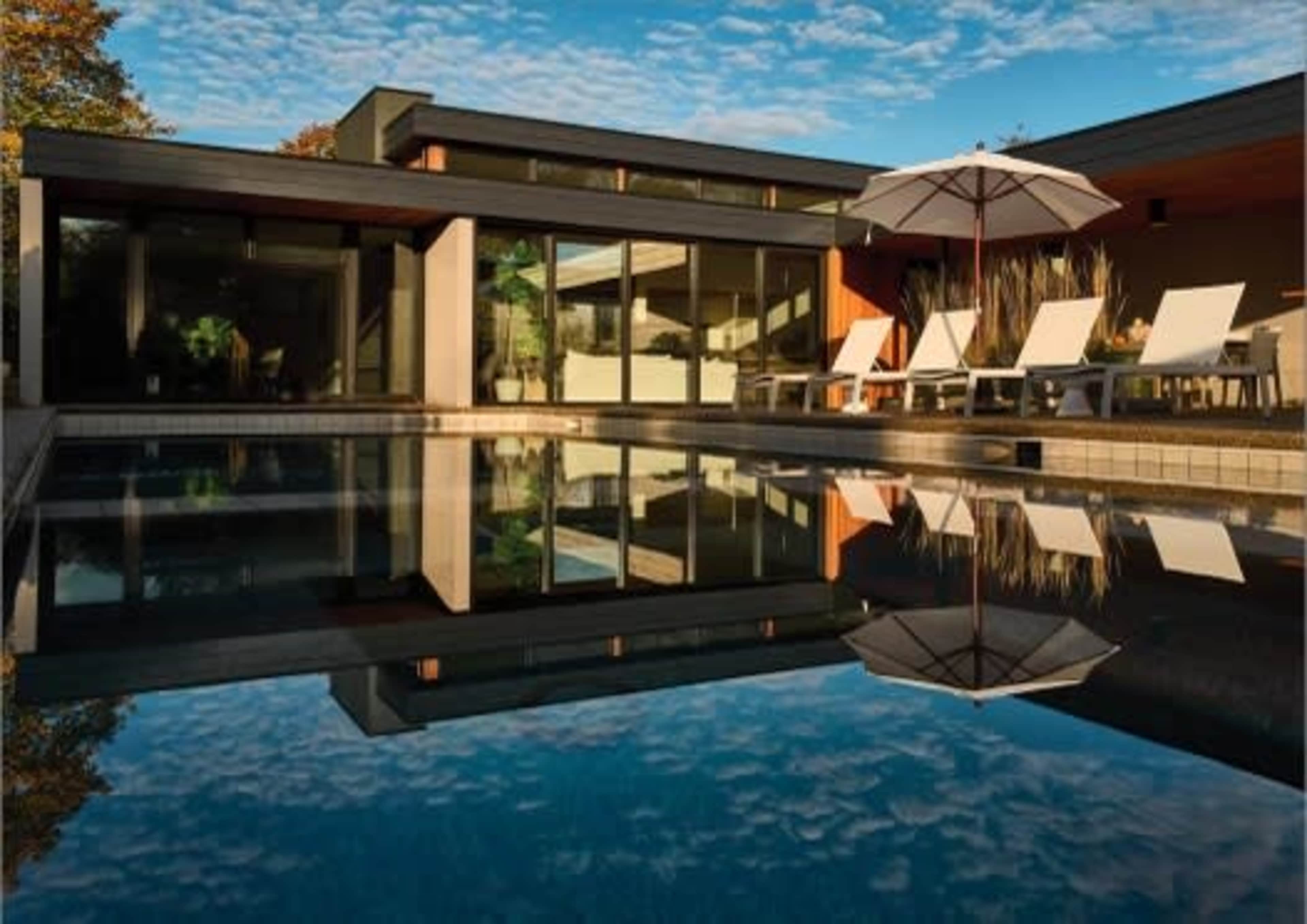 A modern house with large windows overlooks a swimming pool surrounded by lounge chairs and an umbrella, reflecting the sky and trees.