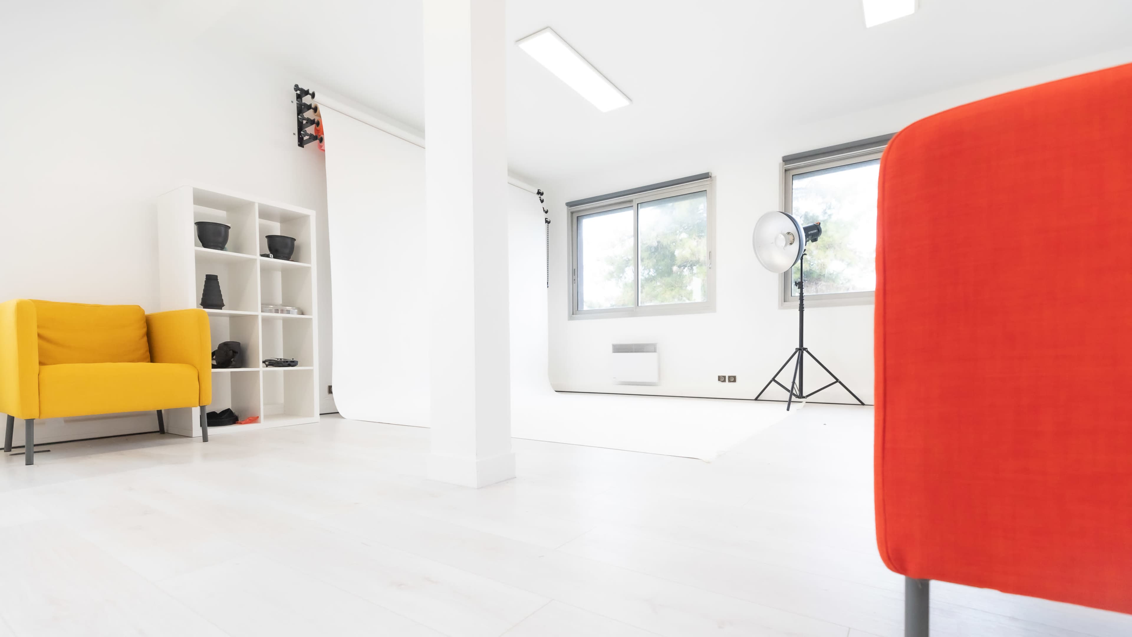 A bright, minimalistic studio space with a yellow armchair, a red chair, a shelving unit, and a large light source near a window.