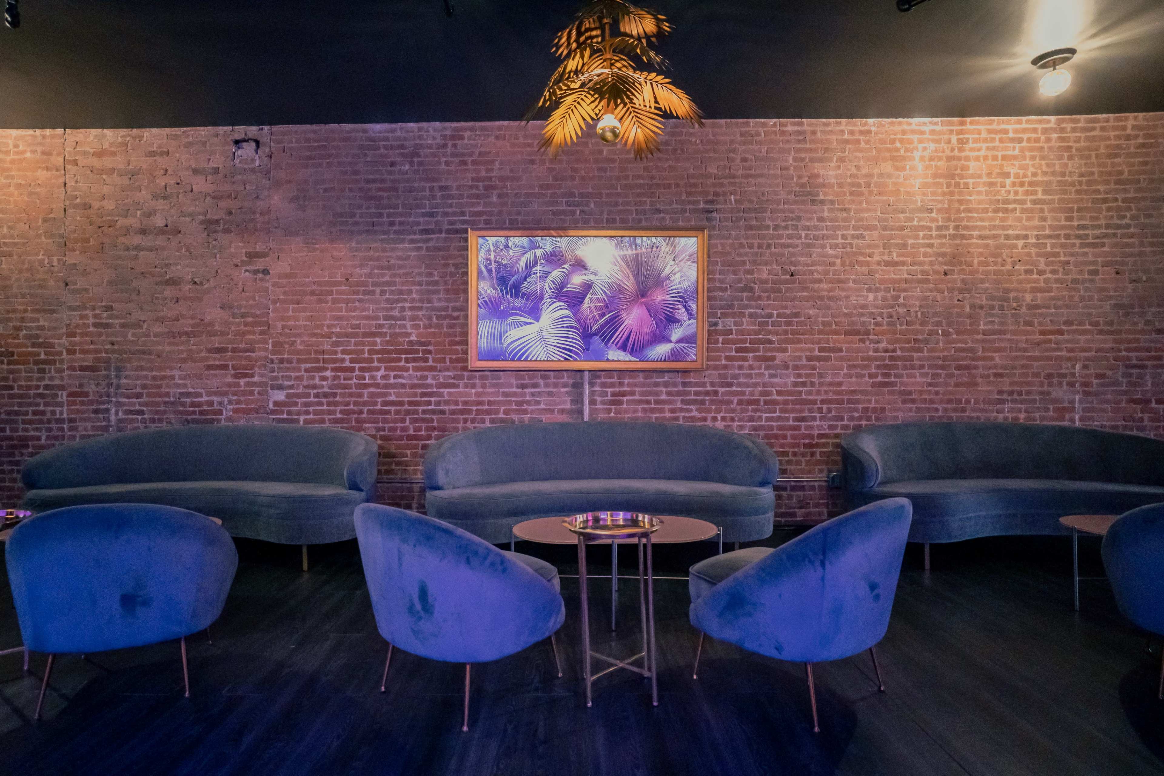 The image shows a dimly lit lounge area with curved, blue velvet sofas, modern chairs, and a framed tropical artwork on a brick wall.