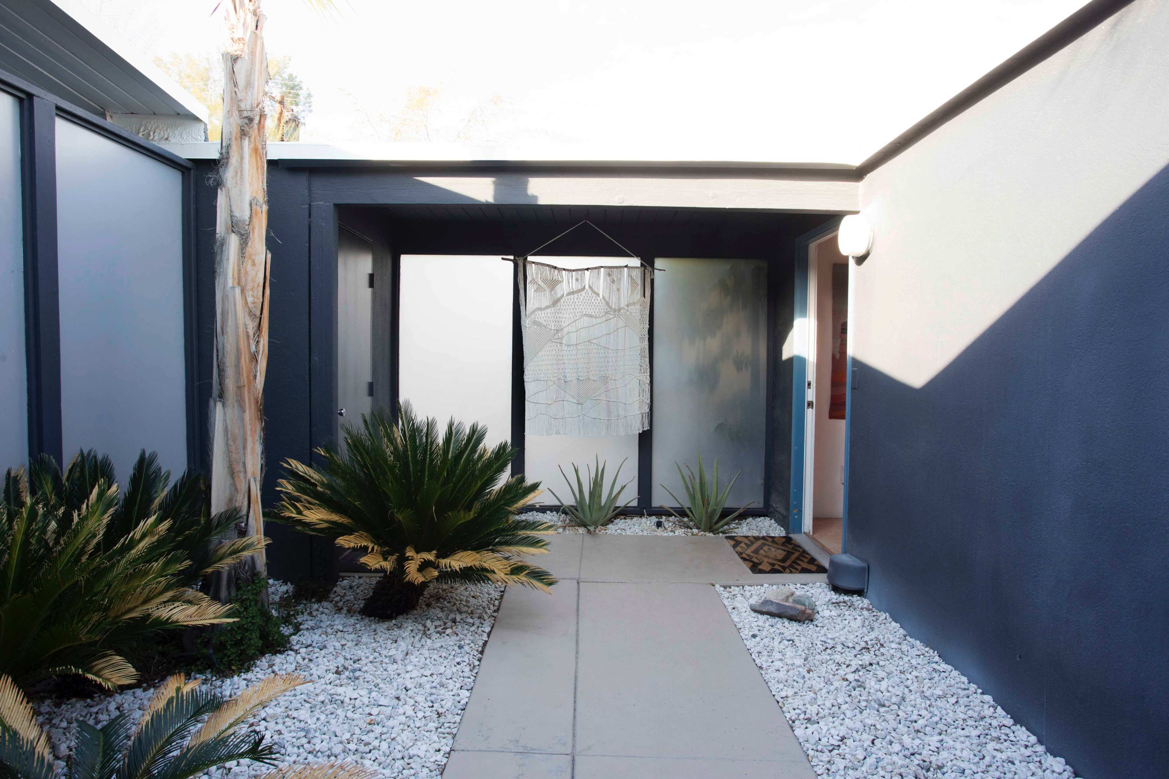A modern entrance pathway flanked by palm plants and white gravel, leading to a doorway with a decorative hanging piece above it.