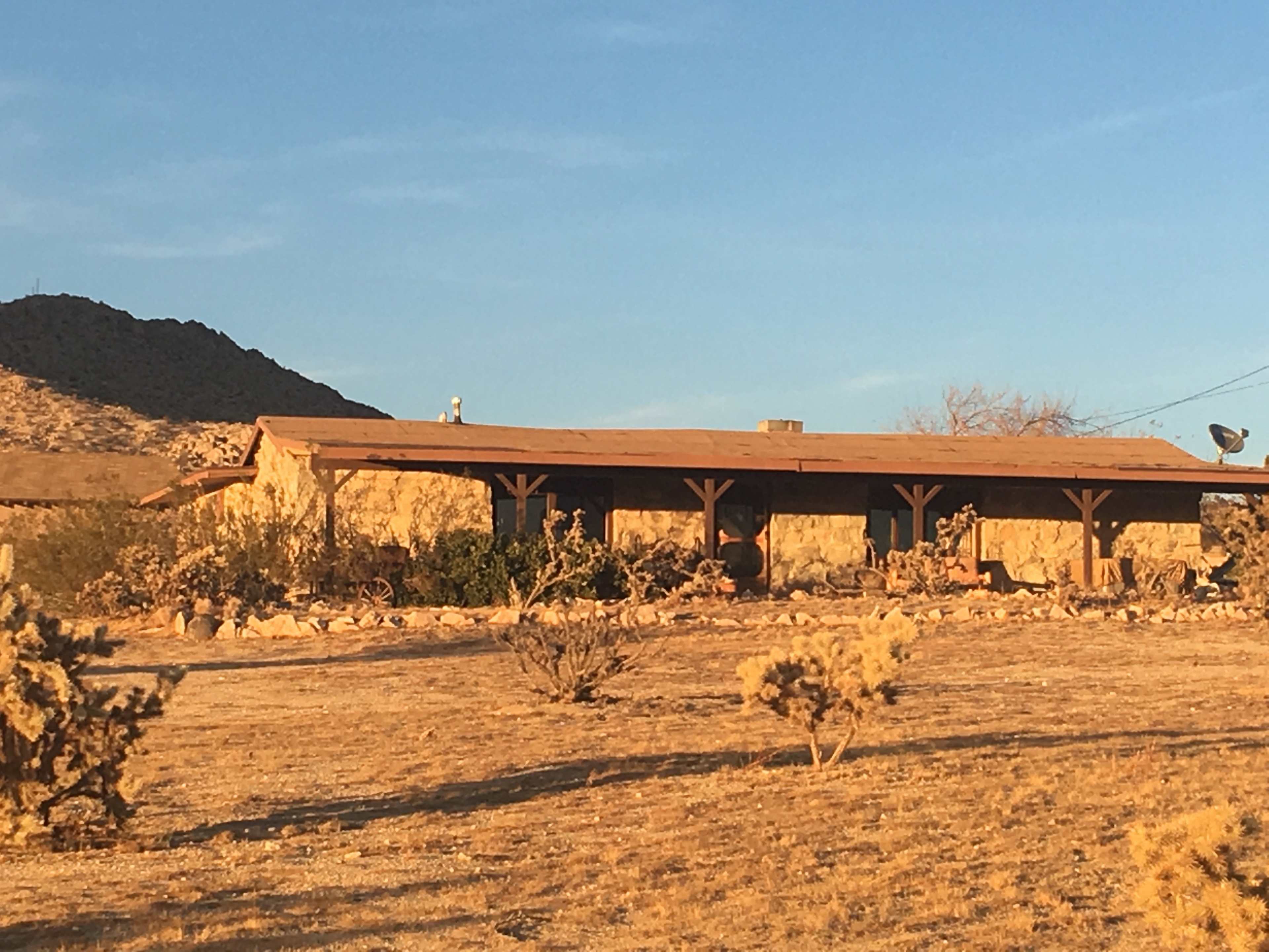 A single-story adobe house stands in a barren landscape with sparse vegetation and distant mountains.