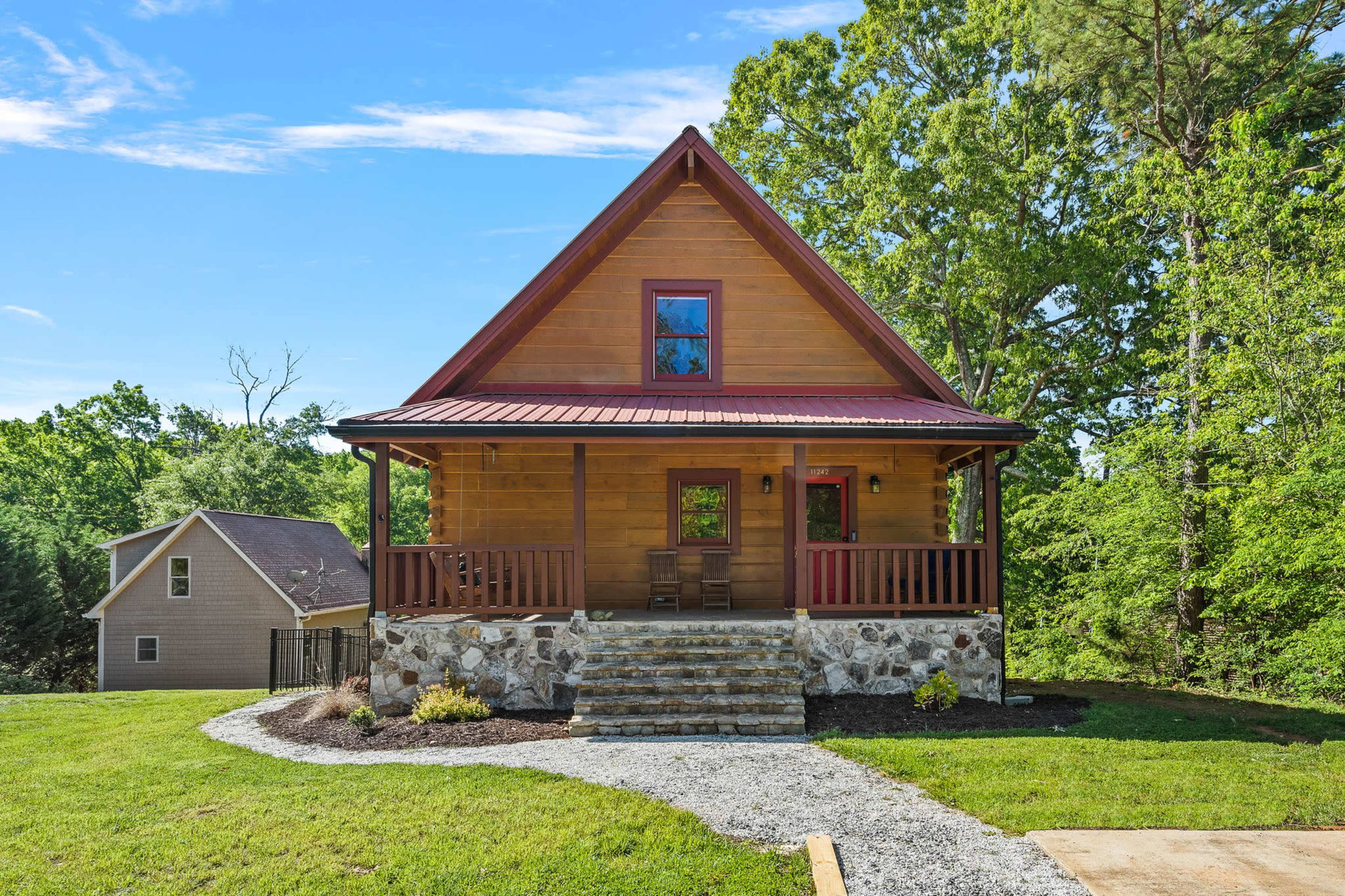 A wooden cabin with a gable roof and stone steps is surrounded by green trees and a grassy yard.