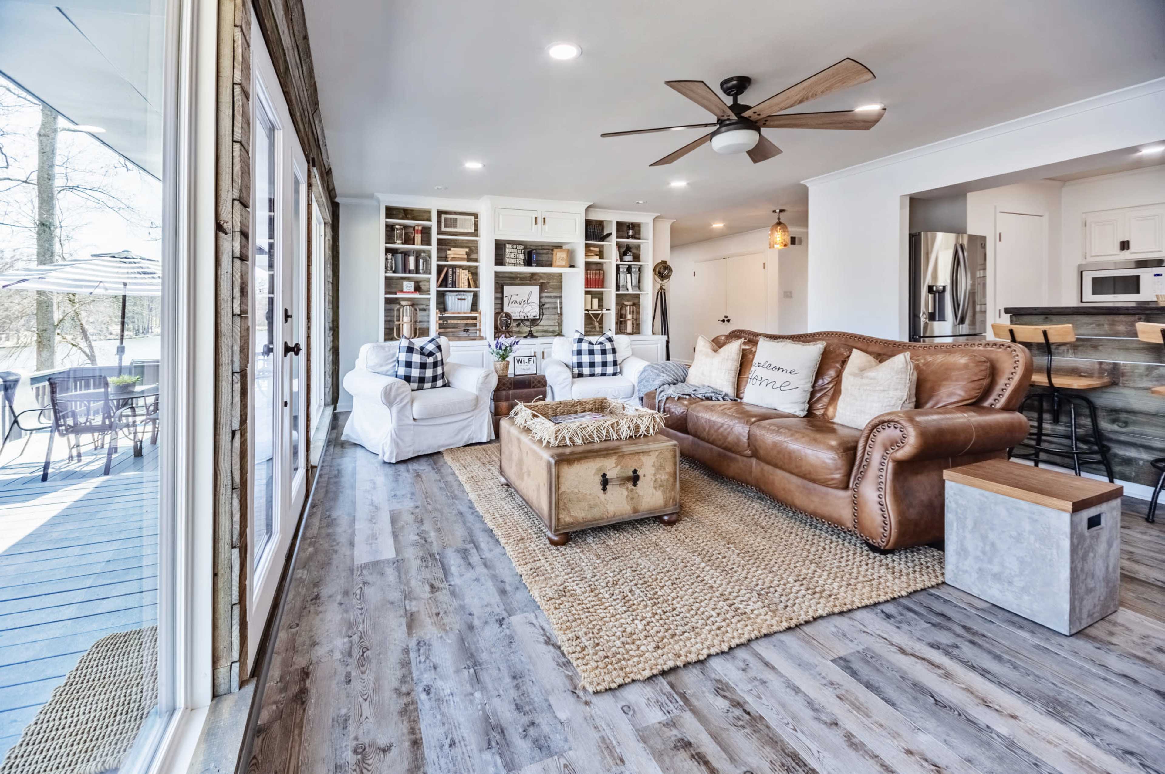 The image shows a bright living room featuring a leather sofa, a wooden coffee table, and bookshelves, with large windows overlooking an outdoor patio.