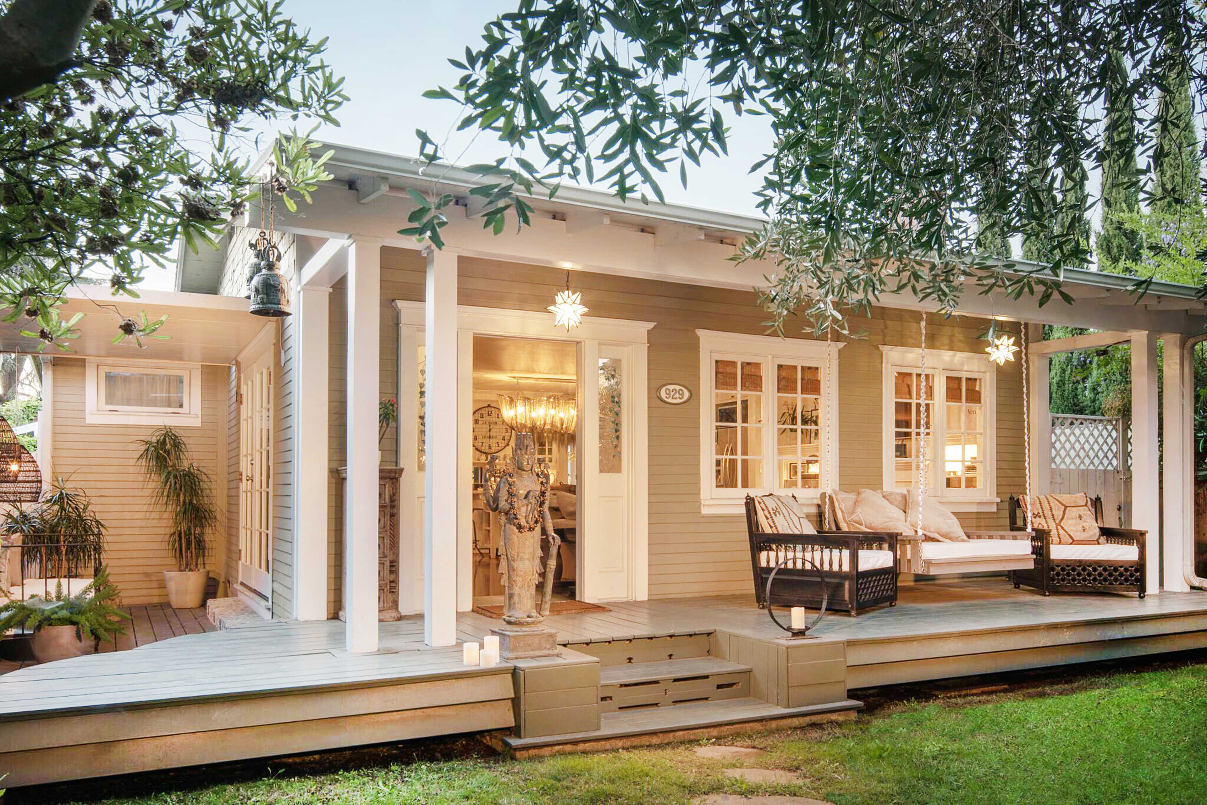 A cozy, light-colored house with a front porch, featuring comfortable seating, surrounded by greenery.