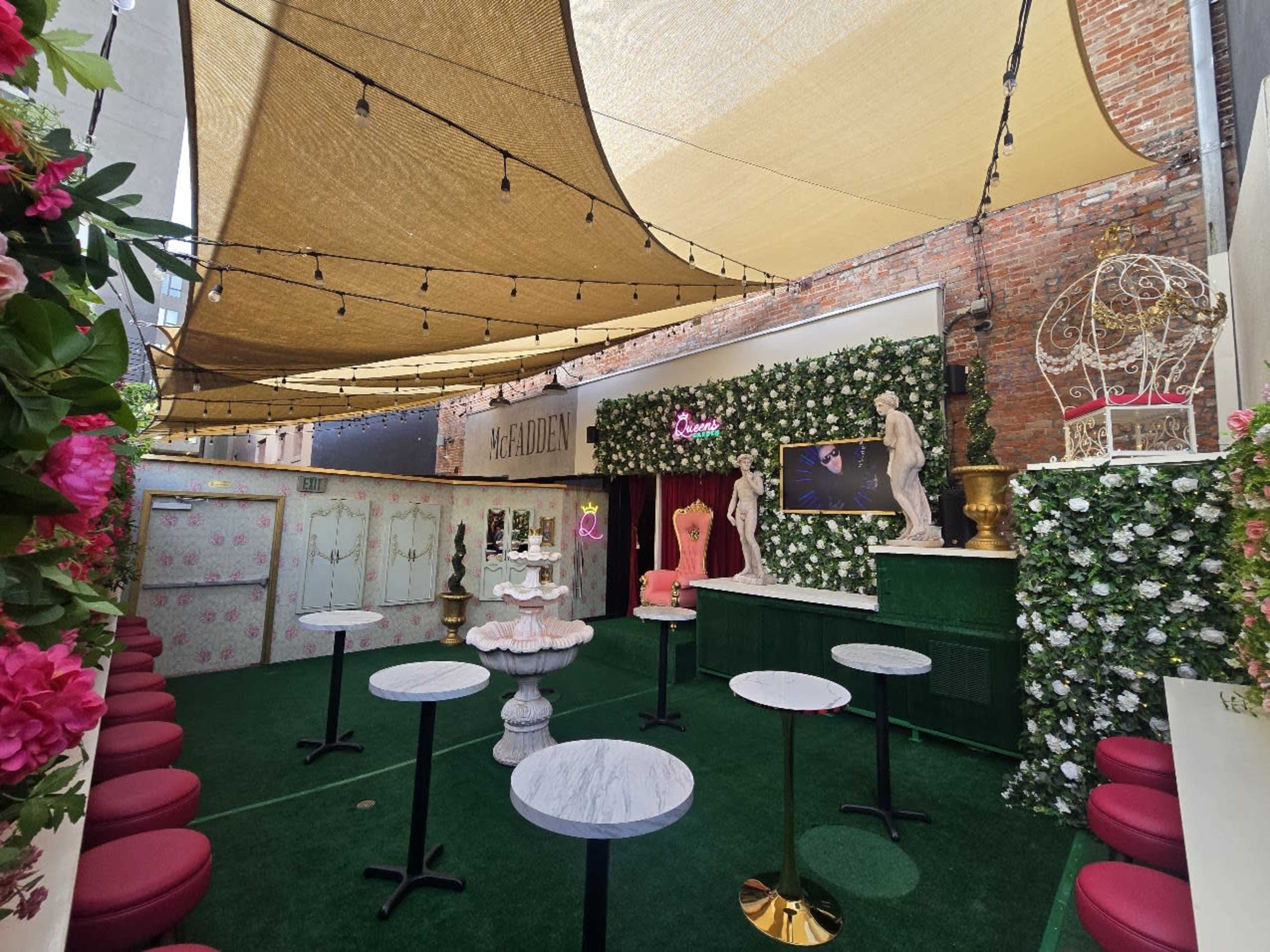 The image shows a decorative outdoor space with green grass, small round tables, and a backdrop of floral walls, complemented by draped fabric overhead for shade.