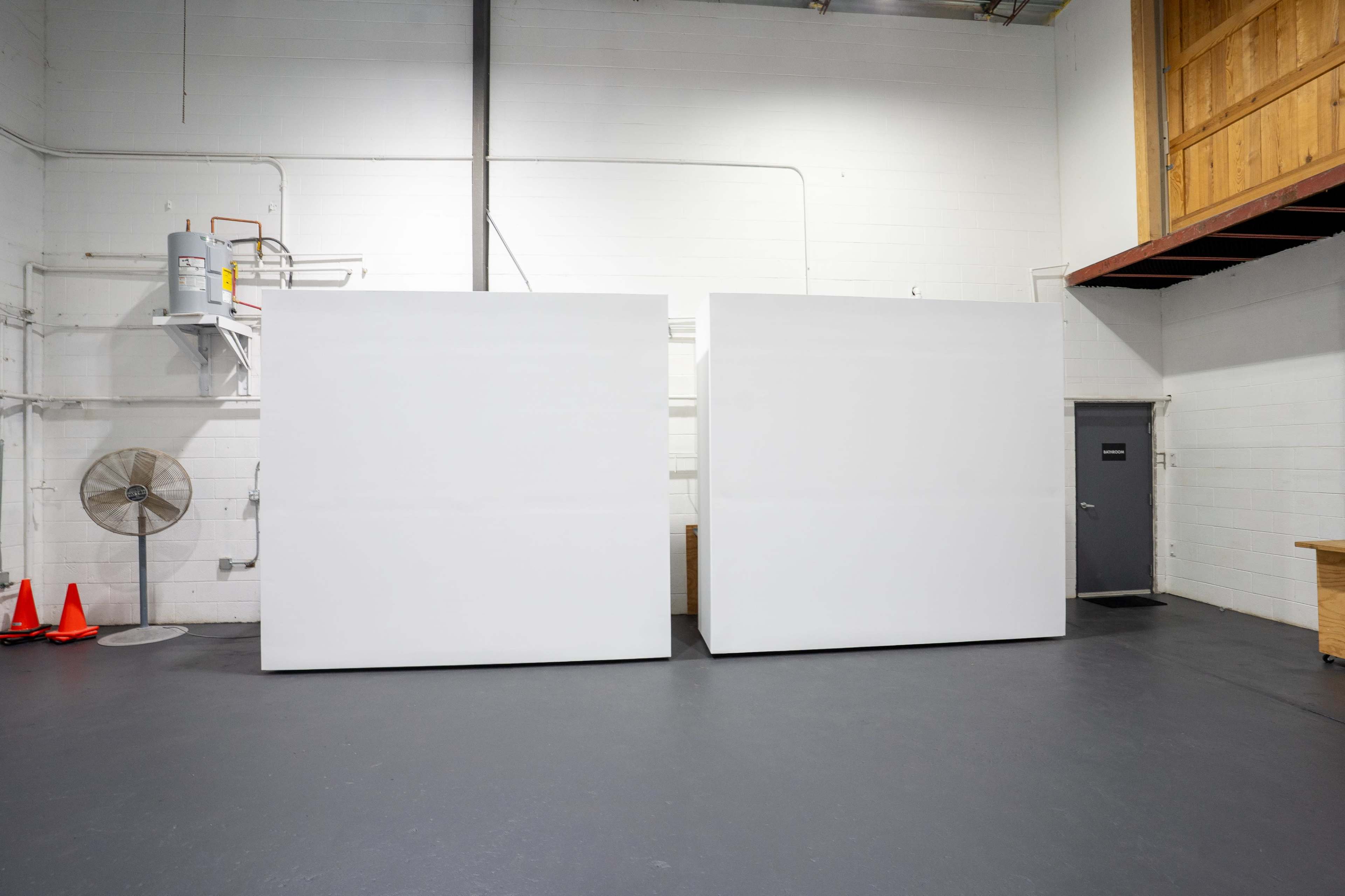 The image shows two large, blank white panels in a warehouse-like space with a concrete floor and industrial features.