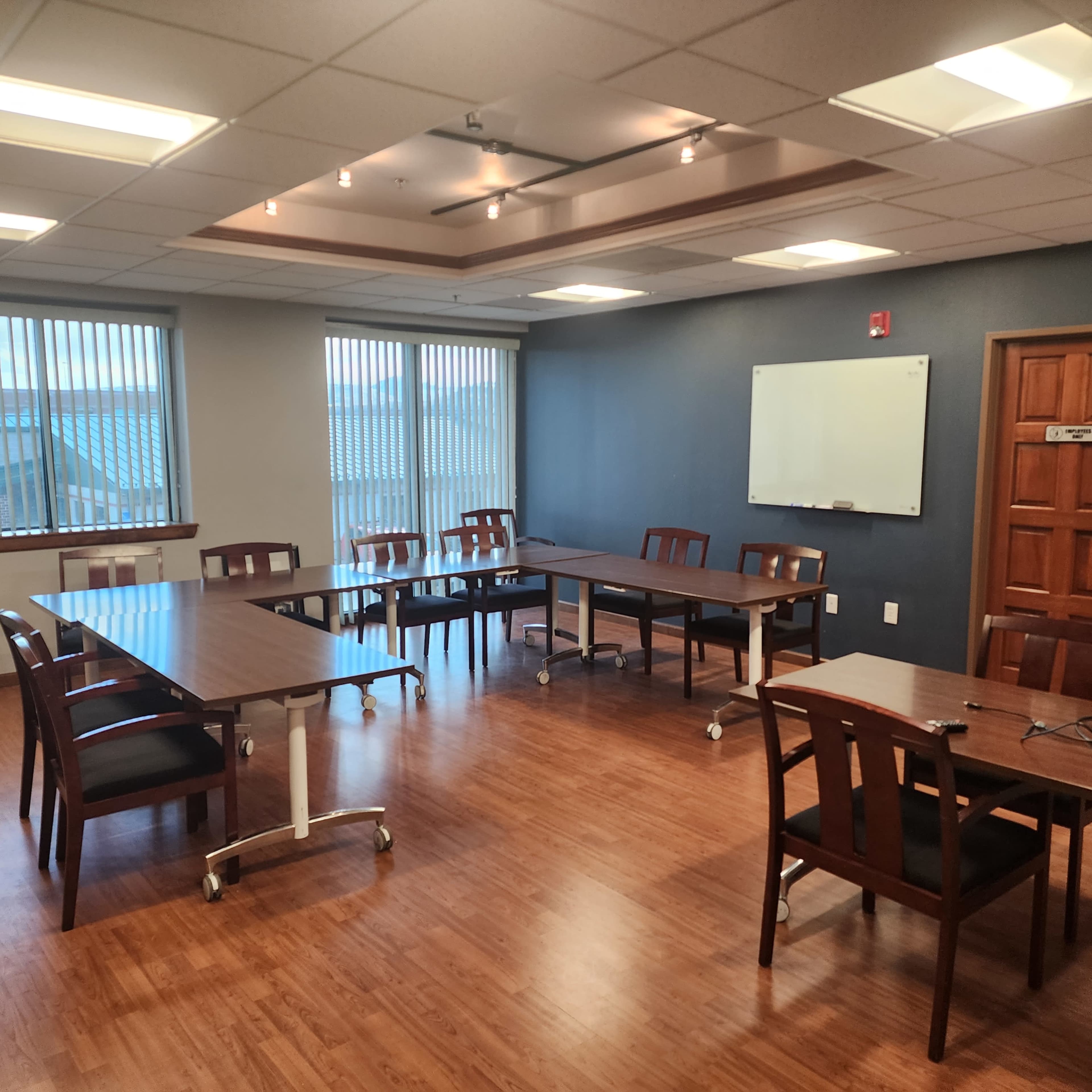 A conference room with multiple tables and chairs arranged around a wooden floor, featuring windows with blinds and a whiteboard on one wall.