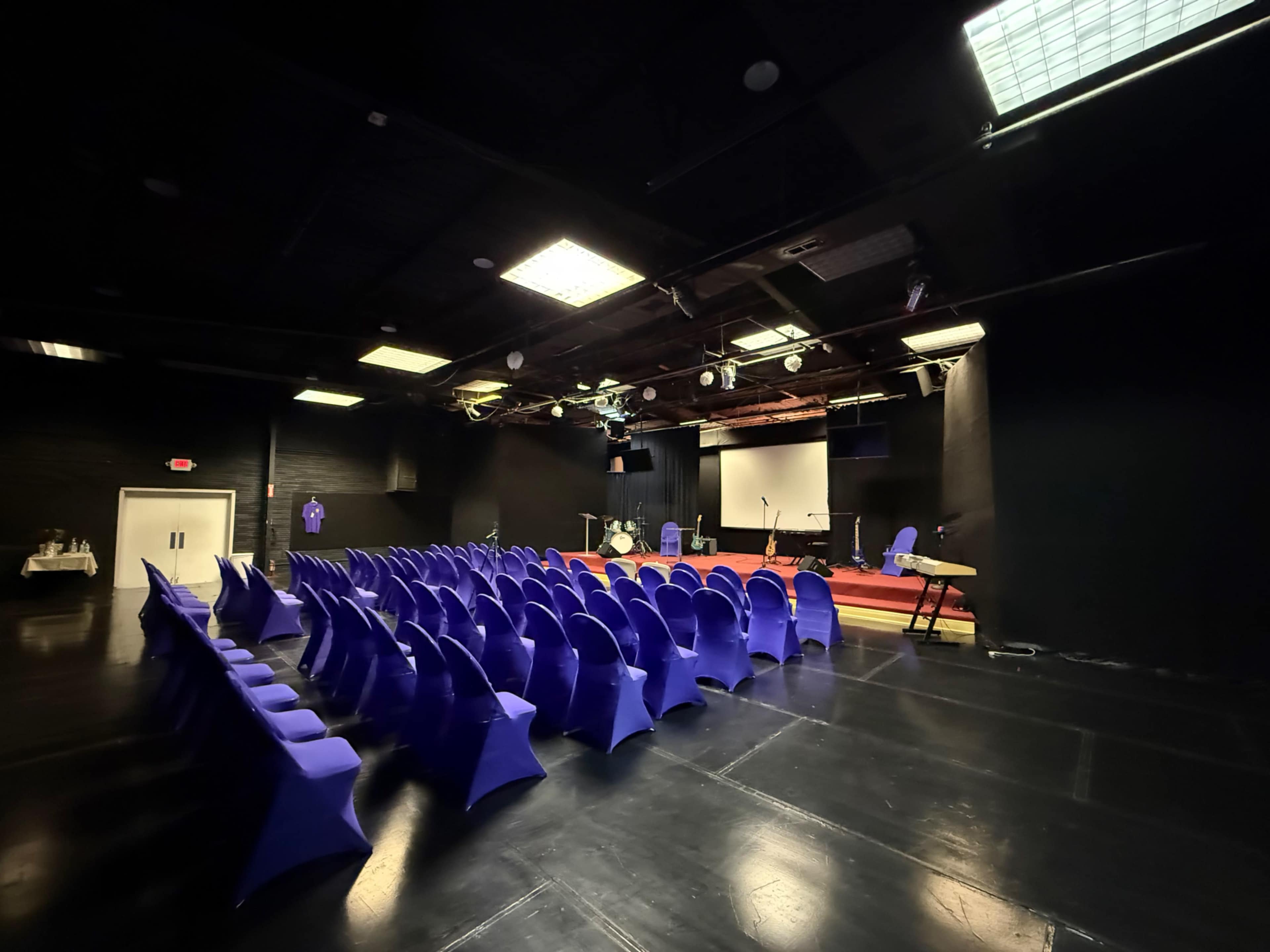 The image depicts an empty auditorium with rows of purple chairs facing a stage that has musical instruments and a large screen.